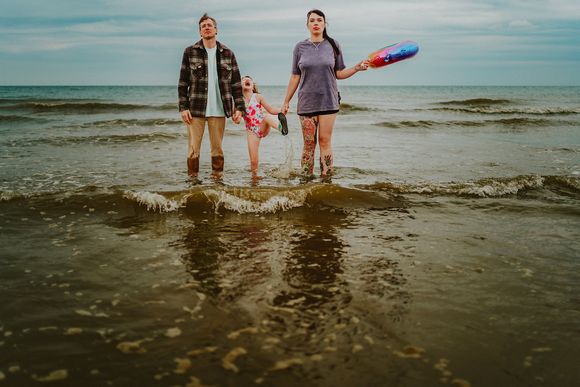 A family stand in the sea at Mablethorpe holding a balloon. The parents stand looking serious while the little girl shouts and splashes.