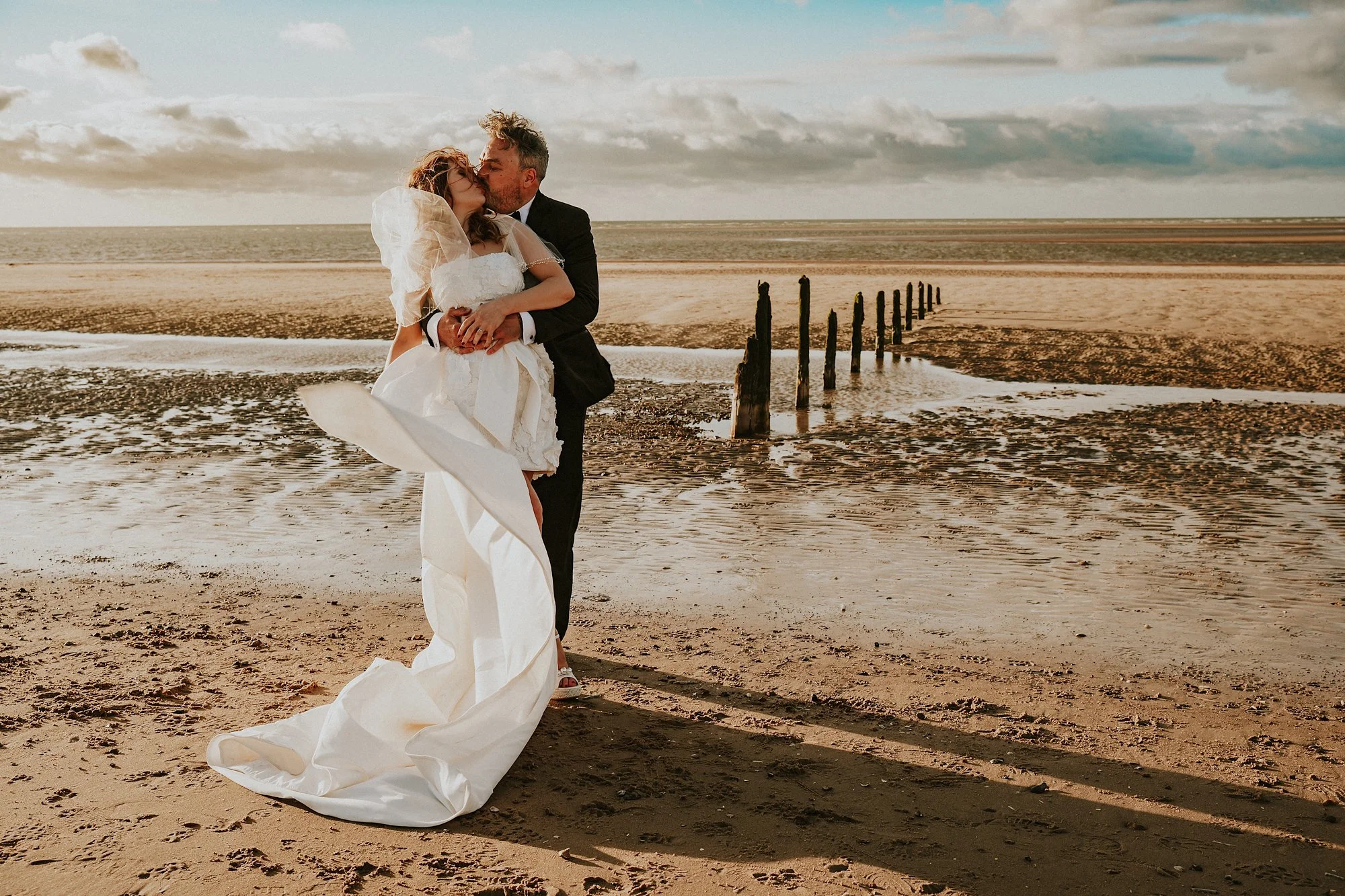 A romantic kiss on the Norfolk Coast as the wind blows the Brides dress. Golden hour and a romantic embrace is the focal point for love in this Titchwell Manor wedding.
