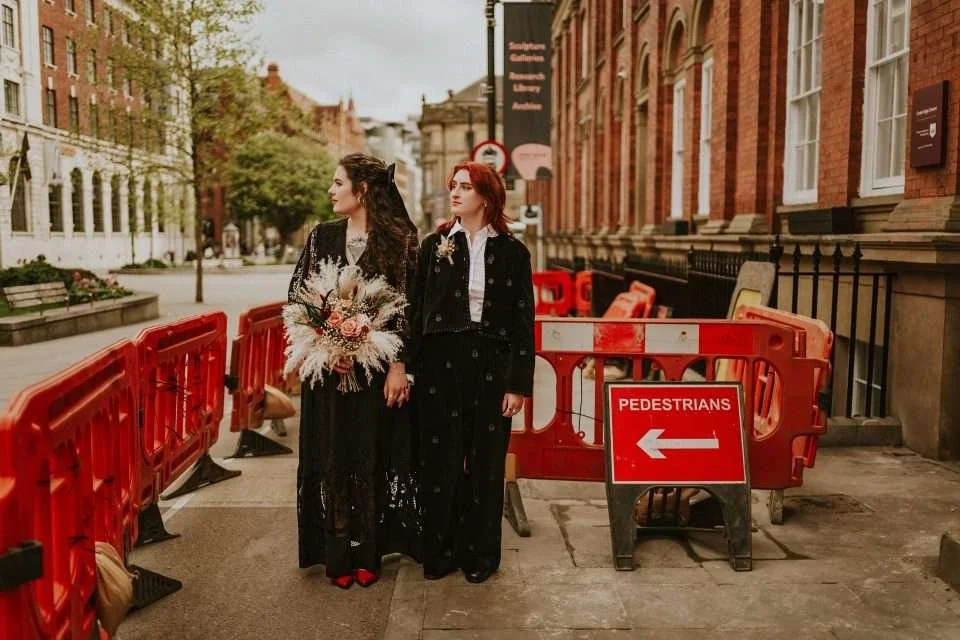 Two women dressed in formal black attire standing on a city sidewalk next to a construction area, with red barriers and a 'PEDESTRIANS' sign with an arrow pointing left. One woman is holding a bouquet of flowers.