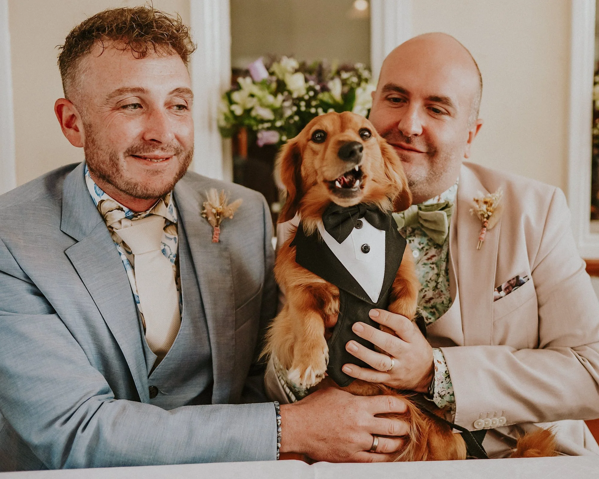 Just married at Bolton Abbey station ticket office. Two grooms smile as they hold their sausage dog in a Tuxedo.