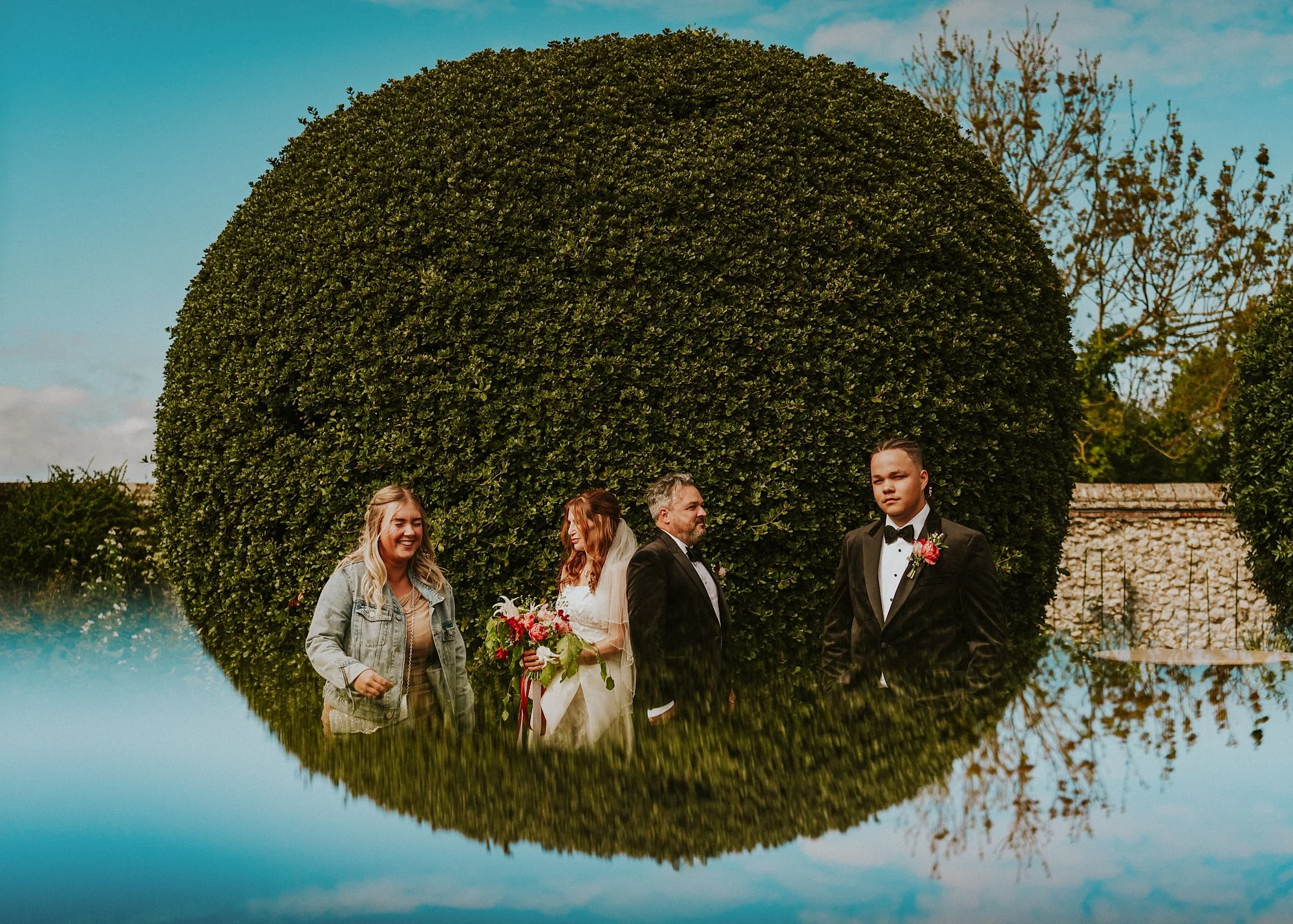 A reflection of a Topiary bush creates a full circle in this quirky group shot of a wedding party at Titchwell Manor.