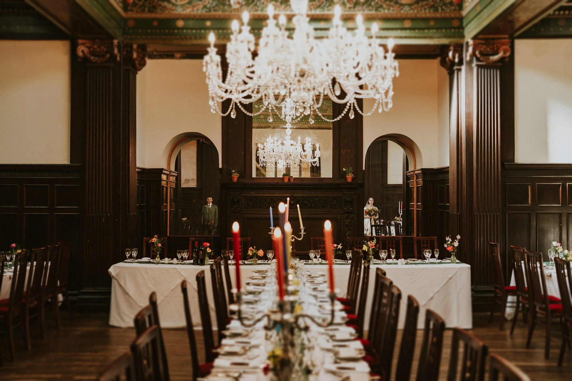 Reflections of the Bride & Groom in Wortley Hall mirrors. Colourful candles, grand chandeliers & dark wood frame an atmospheric portrait.
