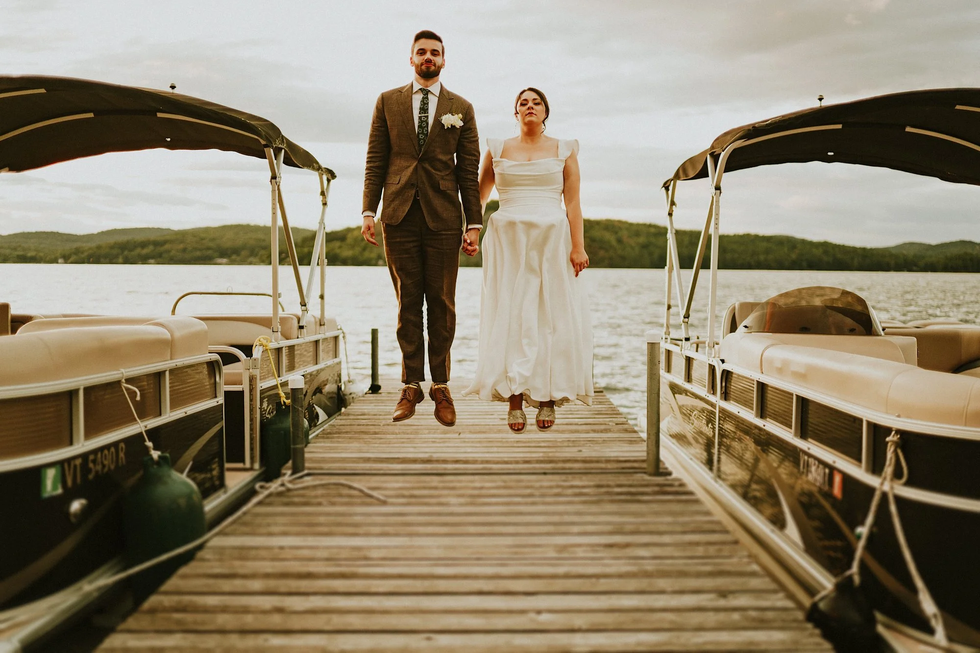 Lake Bomoseen Vermont wedding portrait. A funny image of a couple jumping on the wooden dock surrounded by boats at sunset. Their serious faces and poses make it look like they're floating.