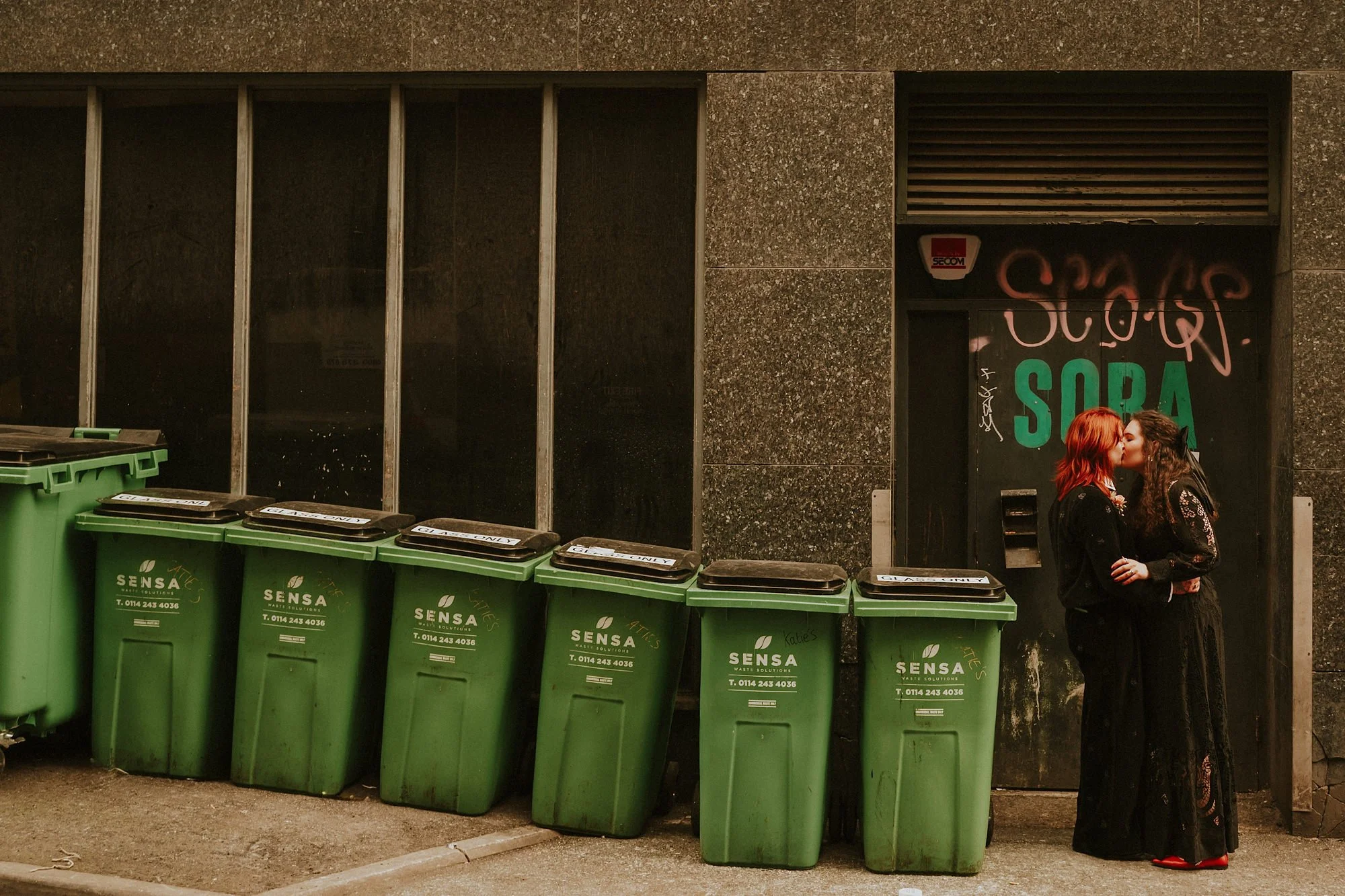 An unusual portrait of two brides in their Leeds wedding with lots of green bins. The couple kiss.