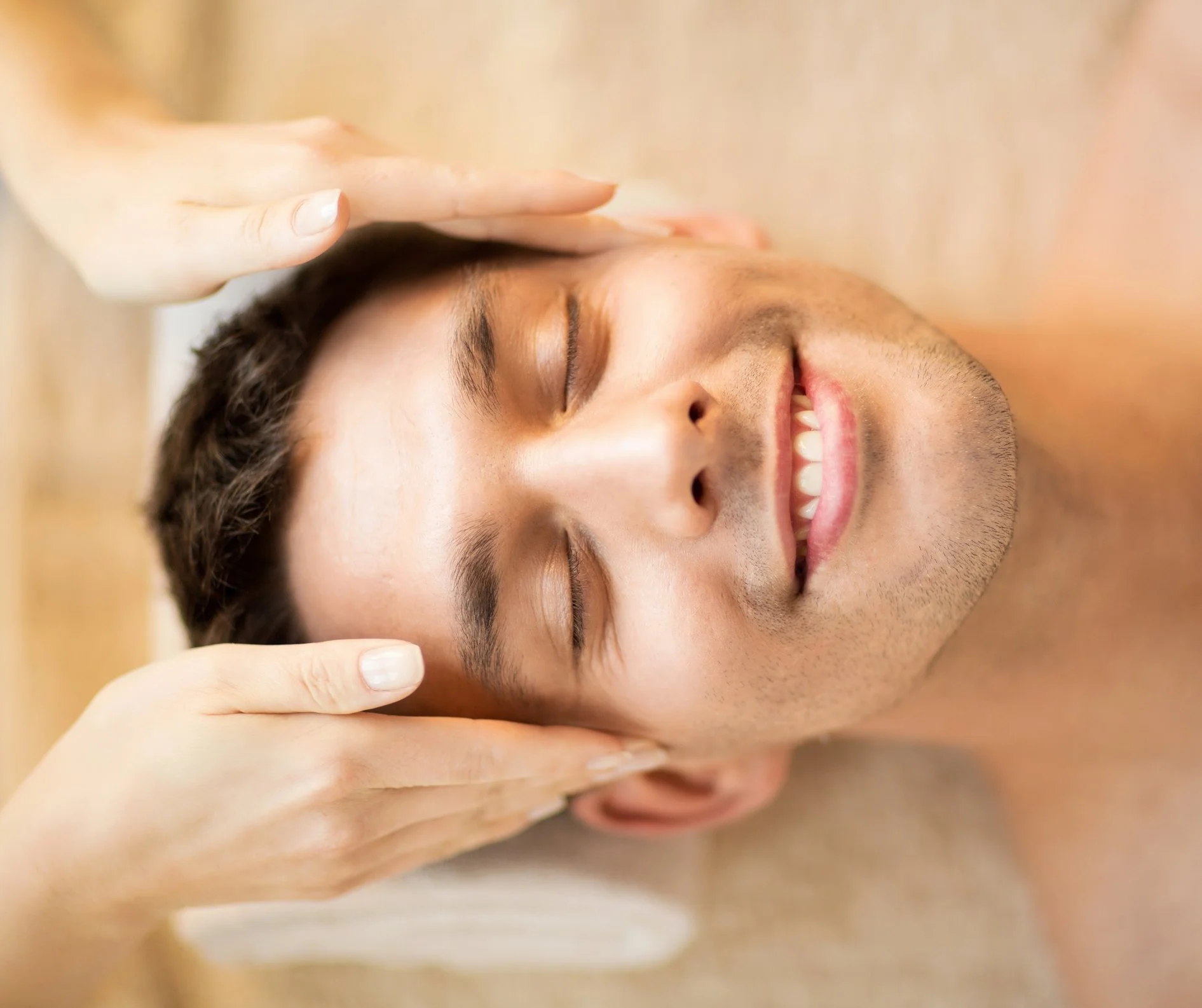 A man smiling with closed eyes, lying down, receiving Reiki from a therapist's hands.