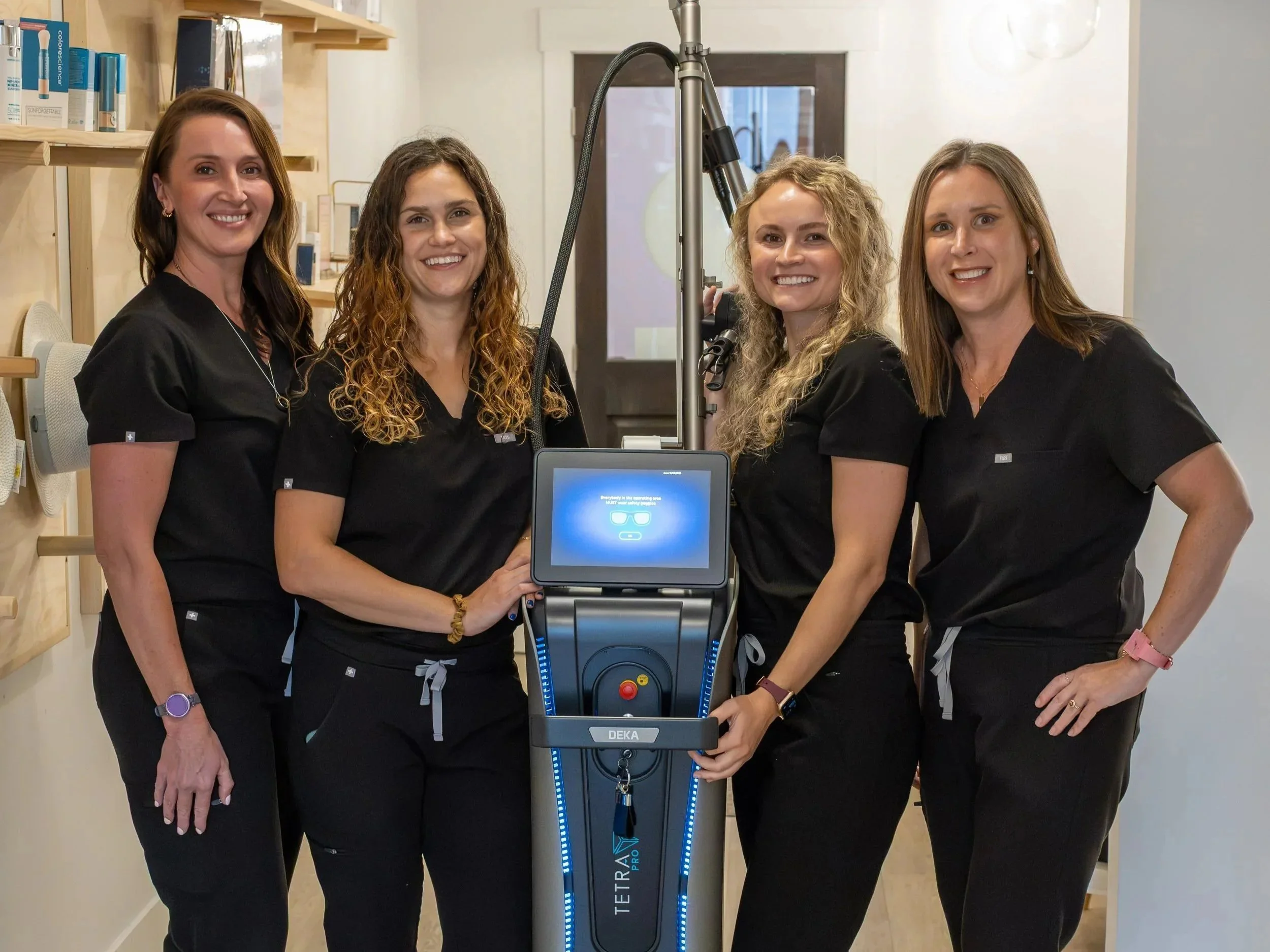 Four women wearing black shirts standing together in a room with shelves and wall decor, posing around a medical device labeled TETRA TRAP PRO CO2 laser with a digital screen and control buttons.