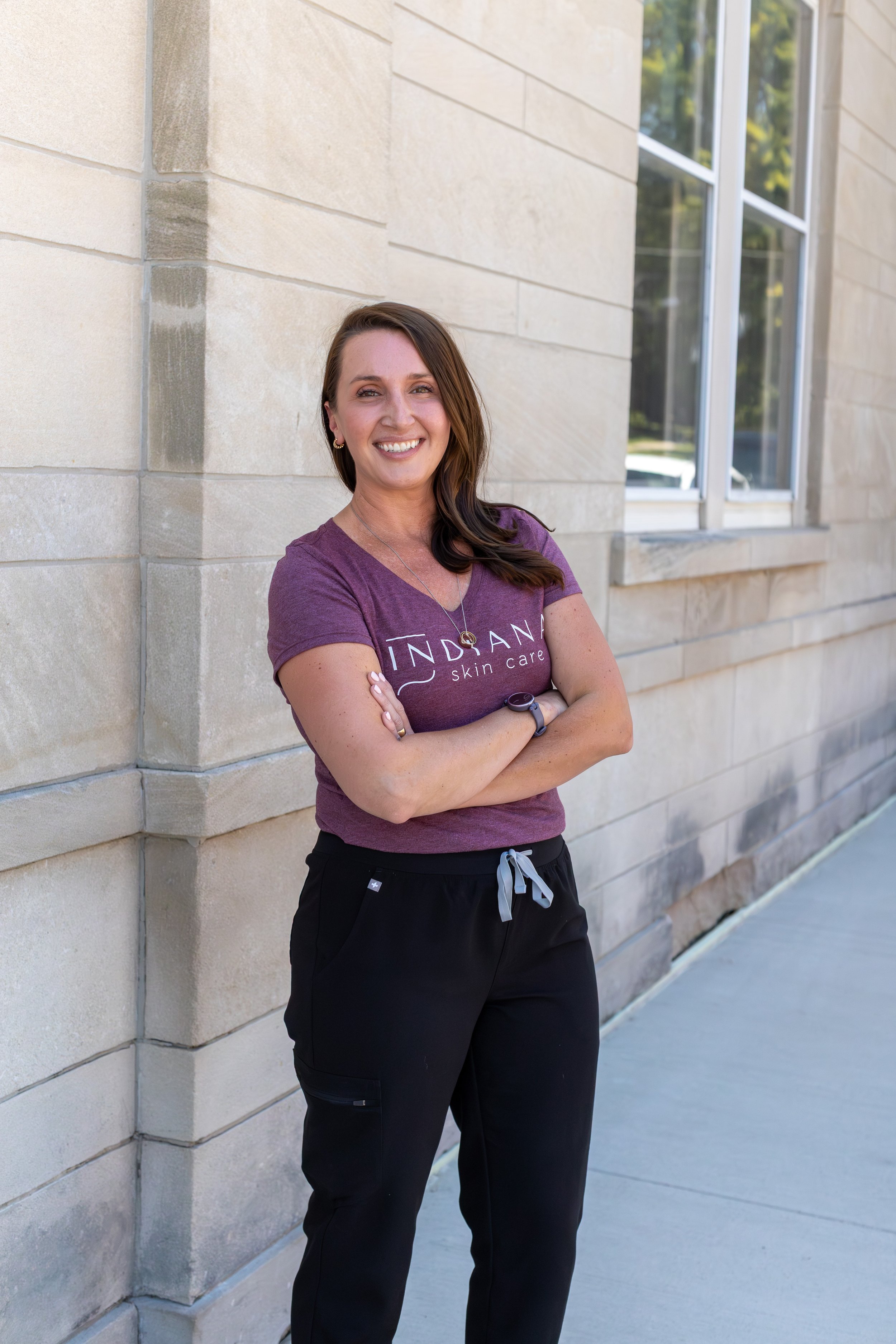 Darcy Hoopingarner smiling with crossed arms, wearing a purple T-shirt and black pants, standing outside against a stone wall and window.