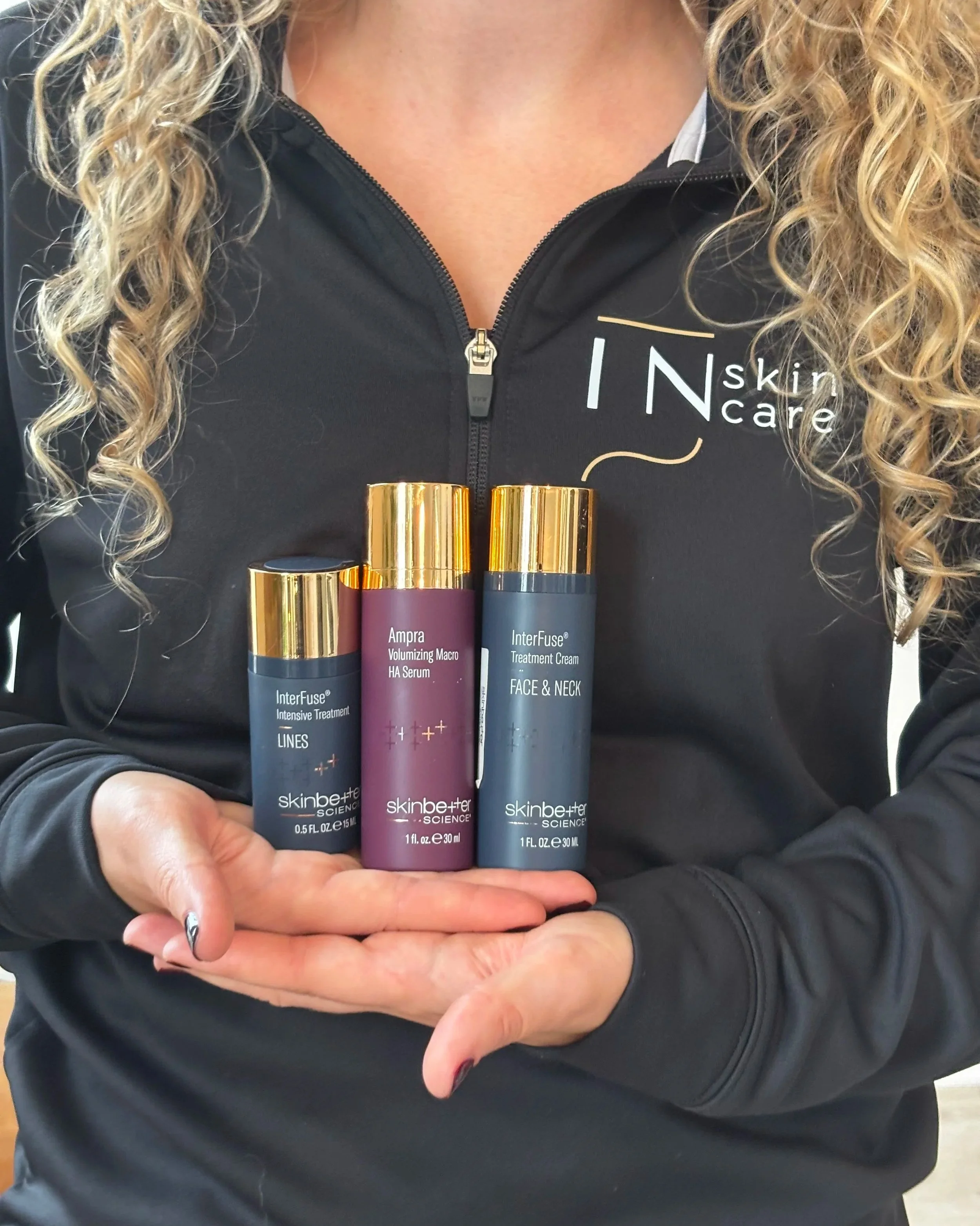 Close-up of a woman wearing a black zip-up jacket with Indiana Skincare company logo, holding three skincare bottles with gold caps.