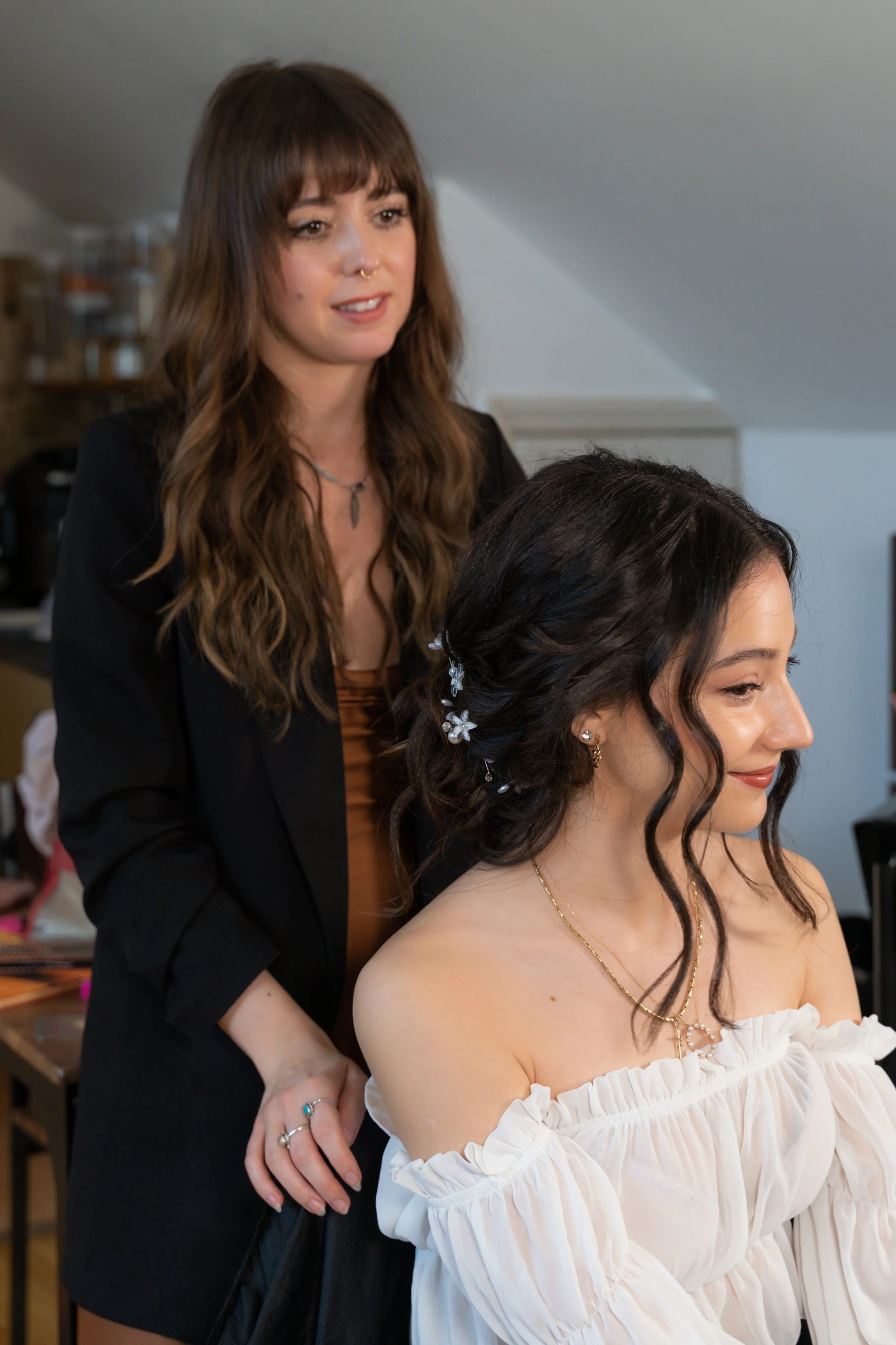 A woman with dark hair styled in loose waves and floral hair accessories is sitting while a hairstylist or makeup artist is working on her, in a room with a slanted ceiling.