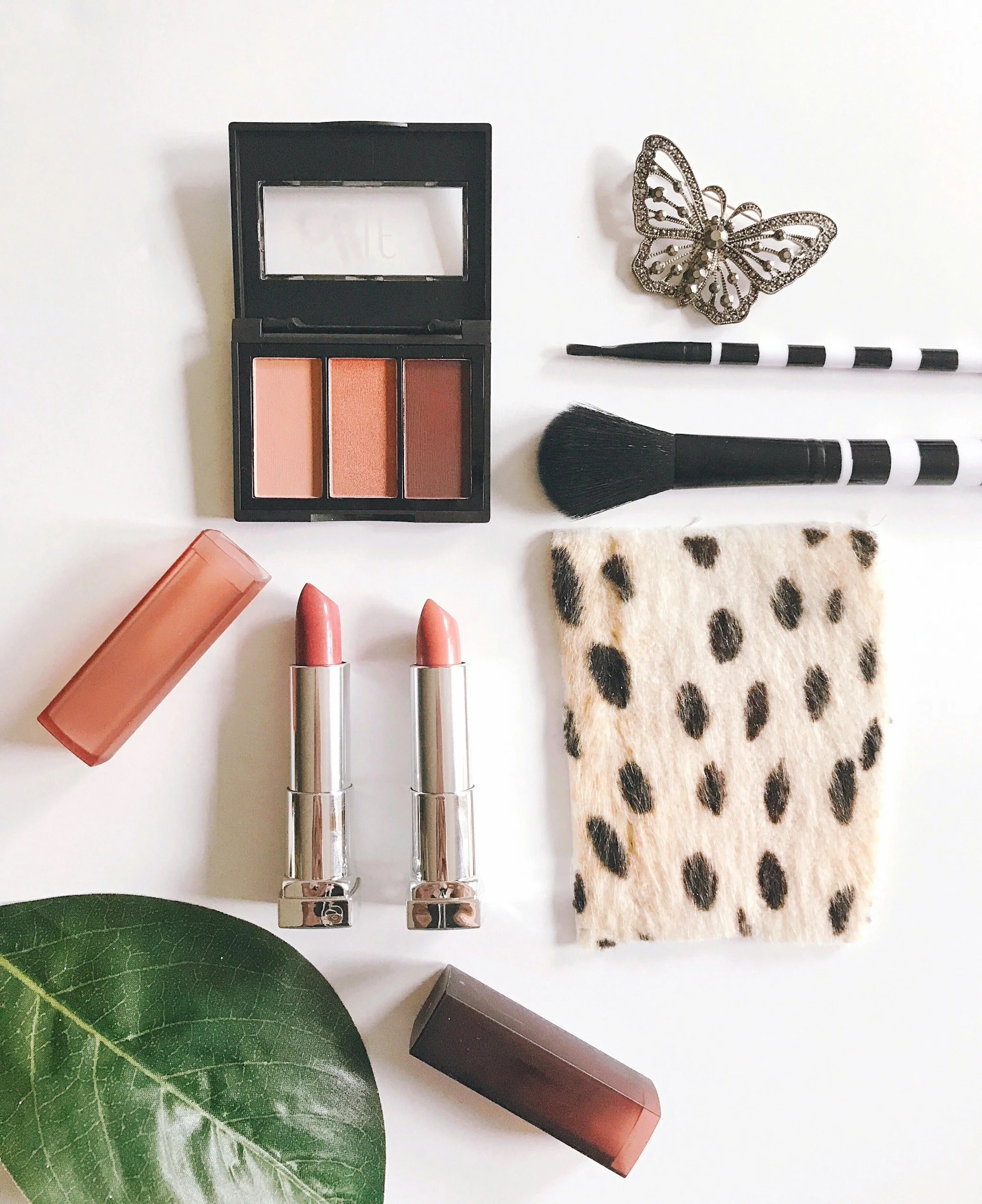 Flat lay of makeup items including a blush palette, two lipsticks, a makeup brush, eye brushes, a butterfly brooch, and a spotted fabric pouch on a white background with a green leaf.