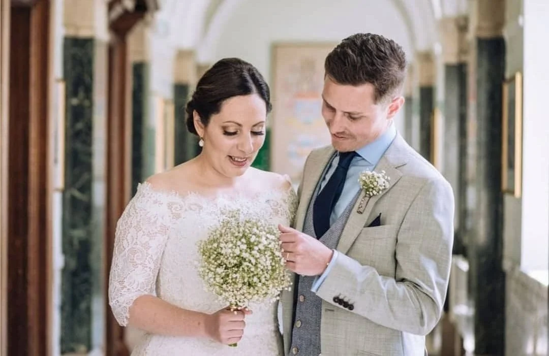 A bride and groom are standing indoors, sharing a moment together. The bride is holding a bouquet, and the groom is touching her left hand, both looking down.
