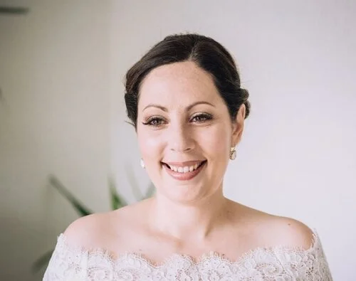 Smiling woman with dark hair in an updo, wearing pearl earrings and a white lace off-the-shoulder top, standing against a light background.