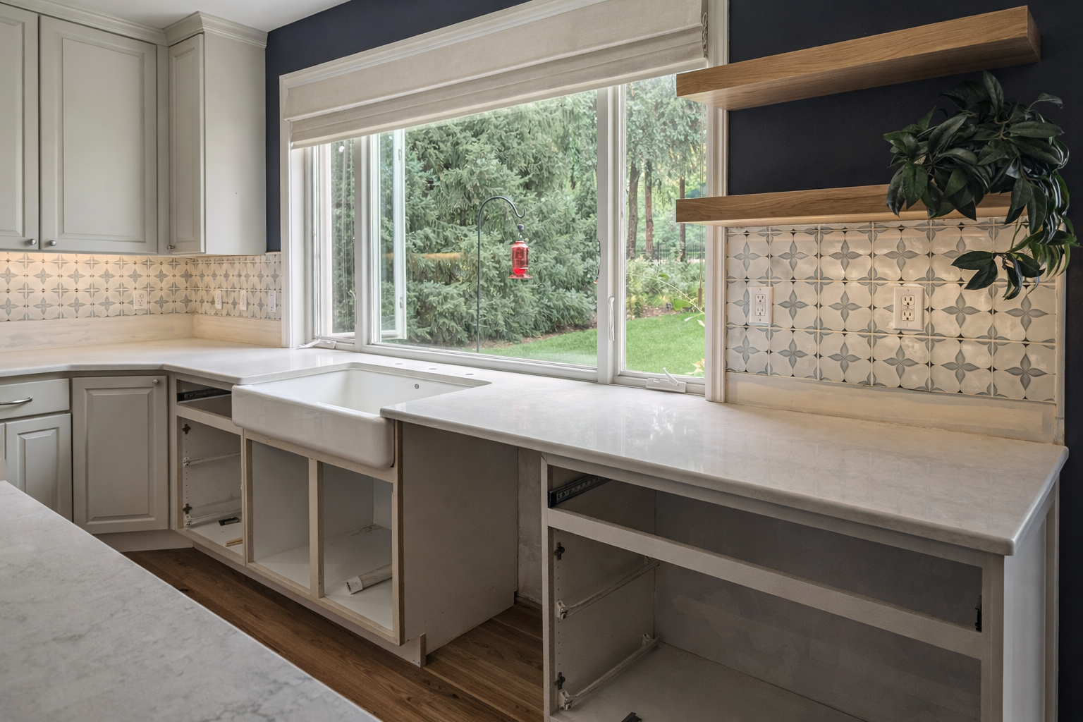 A kitchen under renovation with white cabinets, a large window view of a backyard, a farmhouse sink, and open cabinet space.