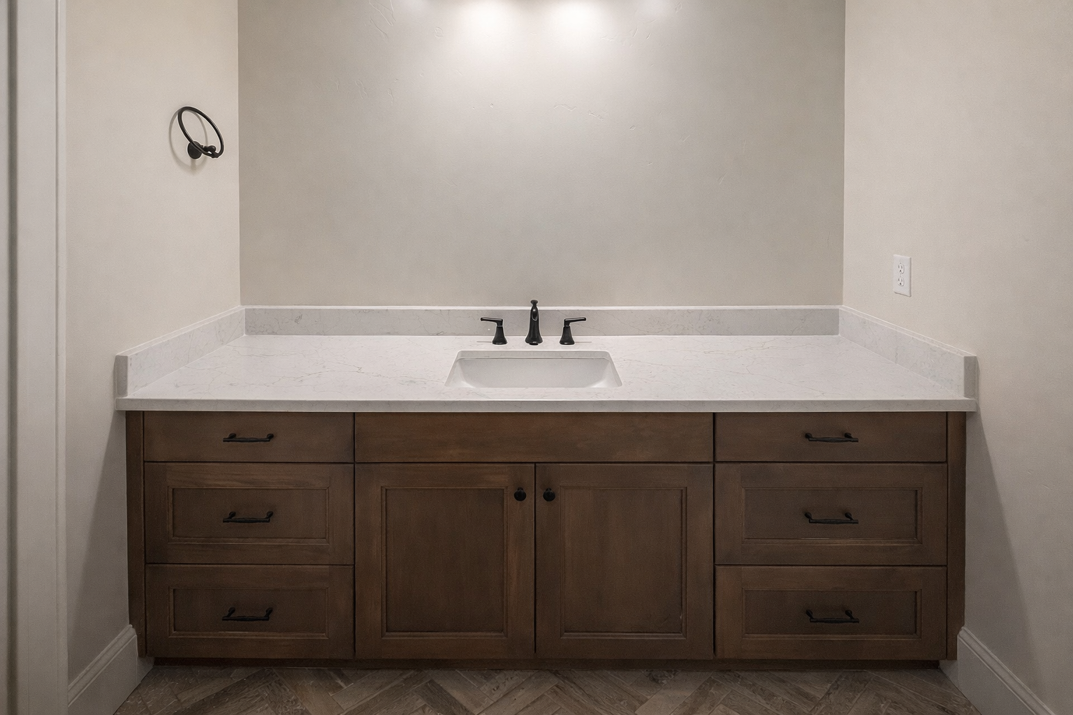 Bathroom vanity with a white marble countertop, a built-in sink, black fixtures, and wooden cabinetry below. There is a wall-mounted towel ring on the left and an electrical outlet on the right wall.