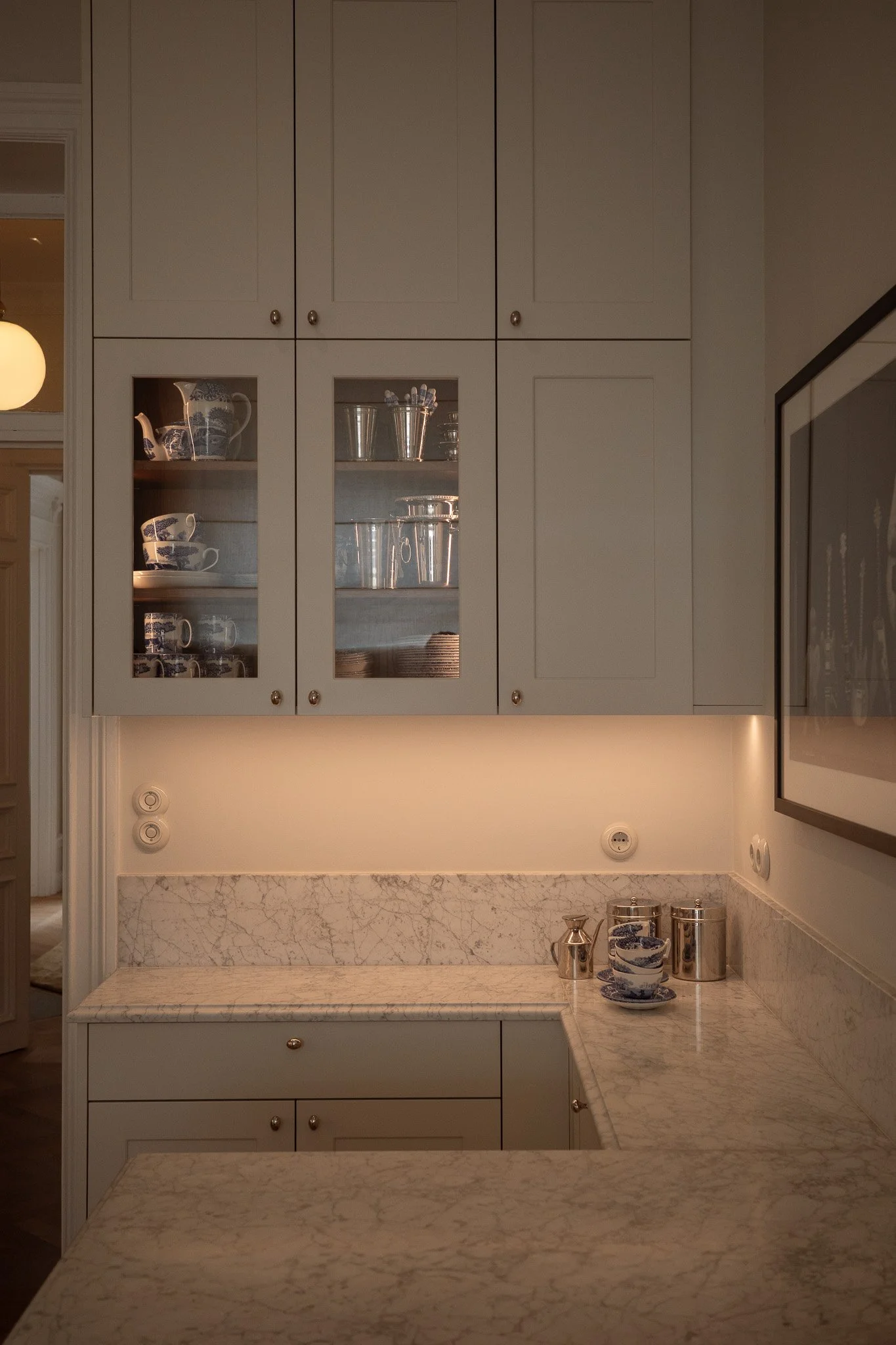 Kitchen with white cabinets, glass-front display with china and silverware, marble countertop, and some kitchen items on the counter.