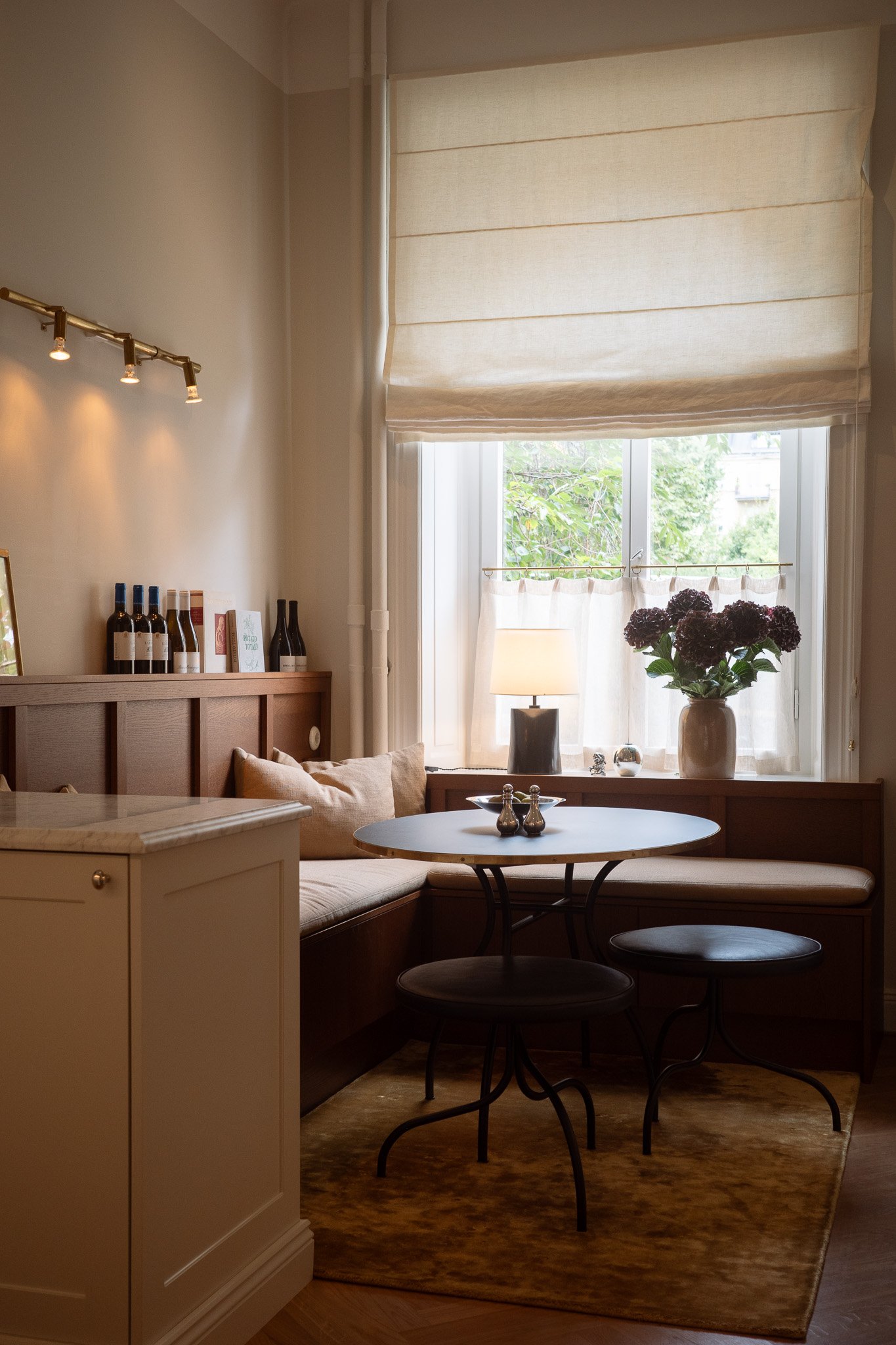 A cozy corner of a living room featuring a window with white curtains and a roman shade, a round table with two black chairs, a large potted purple hydrangea, a beige sofa with cushions, a lamp, and a shelf with wine bottles and framed art.
