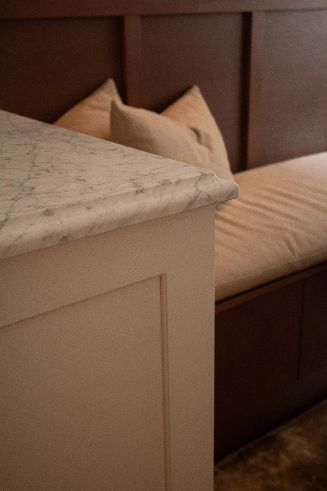 A corner of a nightstand with a marble top next to a bed with beige bedding and pillows, with a wooden headboard in the background.