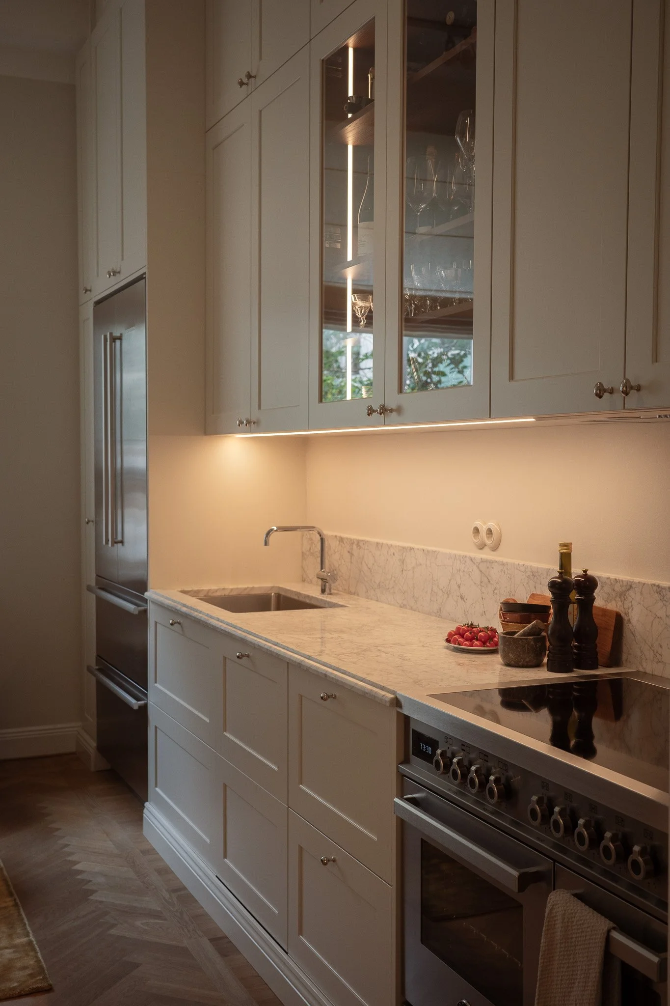 Kitchen with white cabinets, marble countertop, stainless steel stove, and shelf with glassware. Decor includes small bowls, a plate of cherry tomatoes, and salt and pepper shakers. Window with greenery outside reflected in cabinet glass.