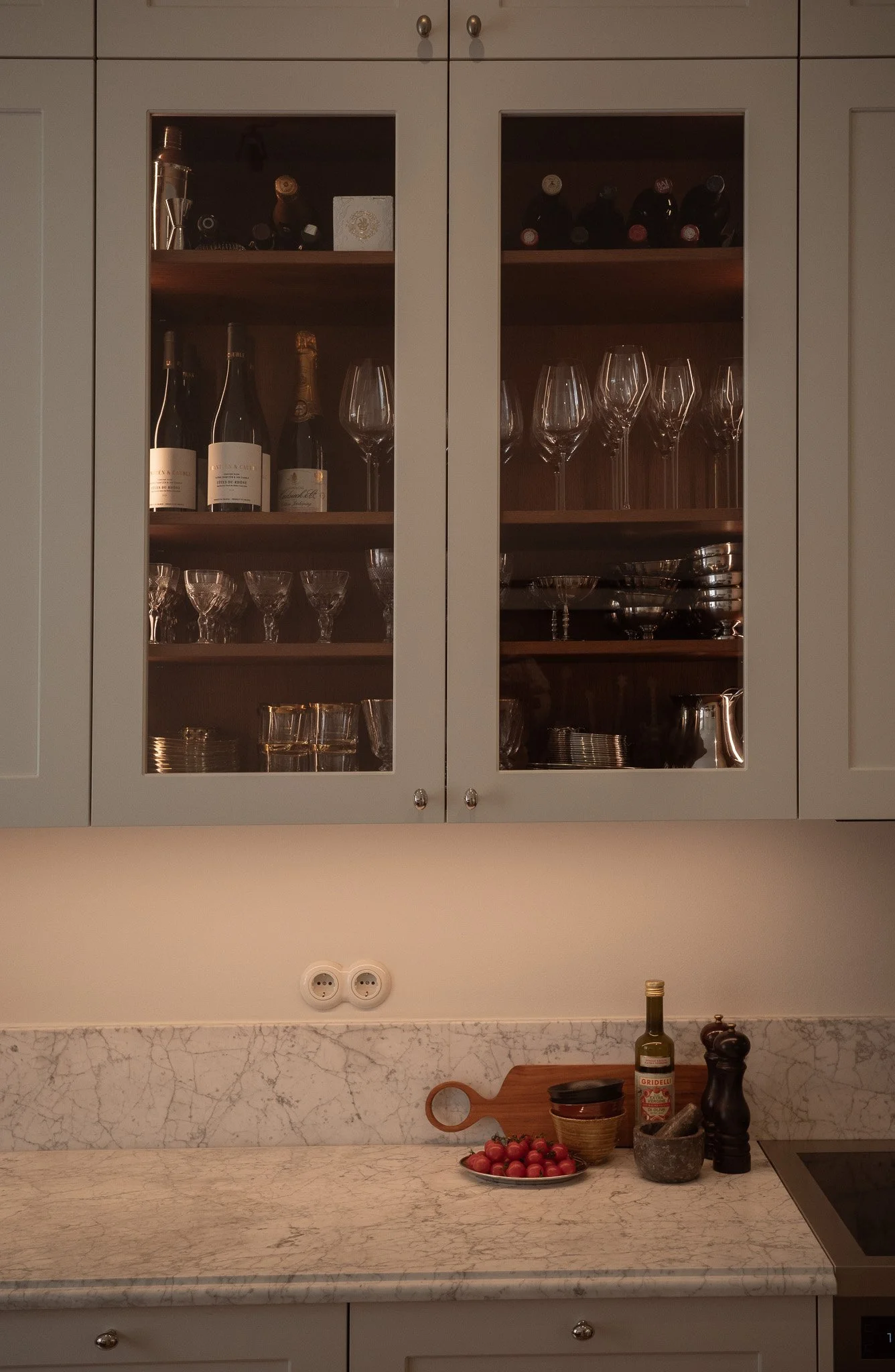 Kitchen cabinet with glass doors containing wine bottles, glasses, and bowls, above a marble countertop with tomatoes, olive oil, pepper grinder, and other kitchen items.