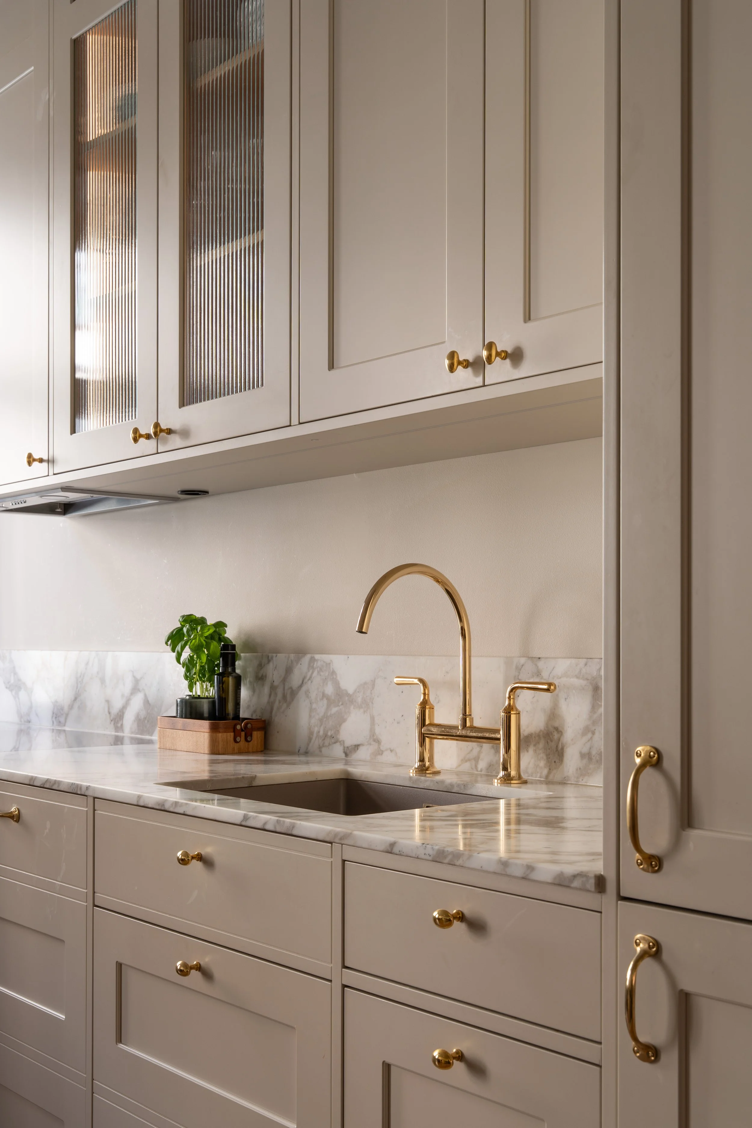 A kitchen with beige cabinets, gold hardware, a marble countertop, and a brass faucet. There is a small green plant and soap dispenser on the counter.