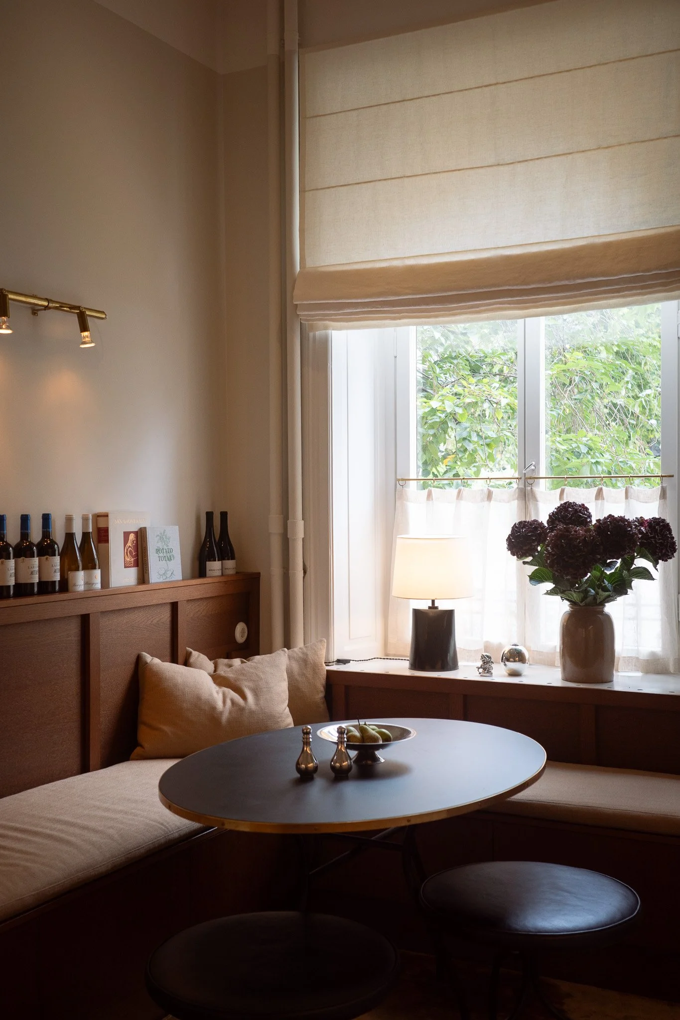 A cozy corner of a room with a window covered by beige Roman shades, a round black table with silver salt and pepper shakers, a bowl of pears, a neutral-colored cushioned bench with pillows, a vase with dark purple hydrangeas, a lamp, and a bookshelf