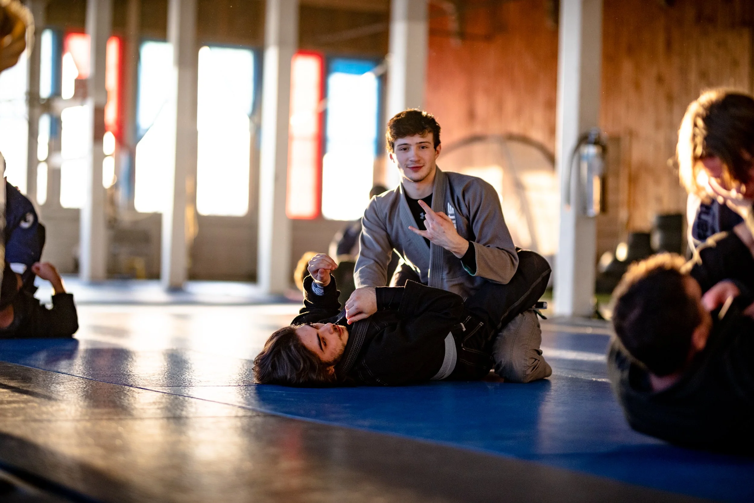 Two young men engaged in a friendly grappling match on a blue martial arts mat in a brightly lit indoor gym with wooden walls and colorful windows.
