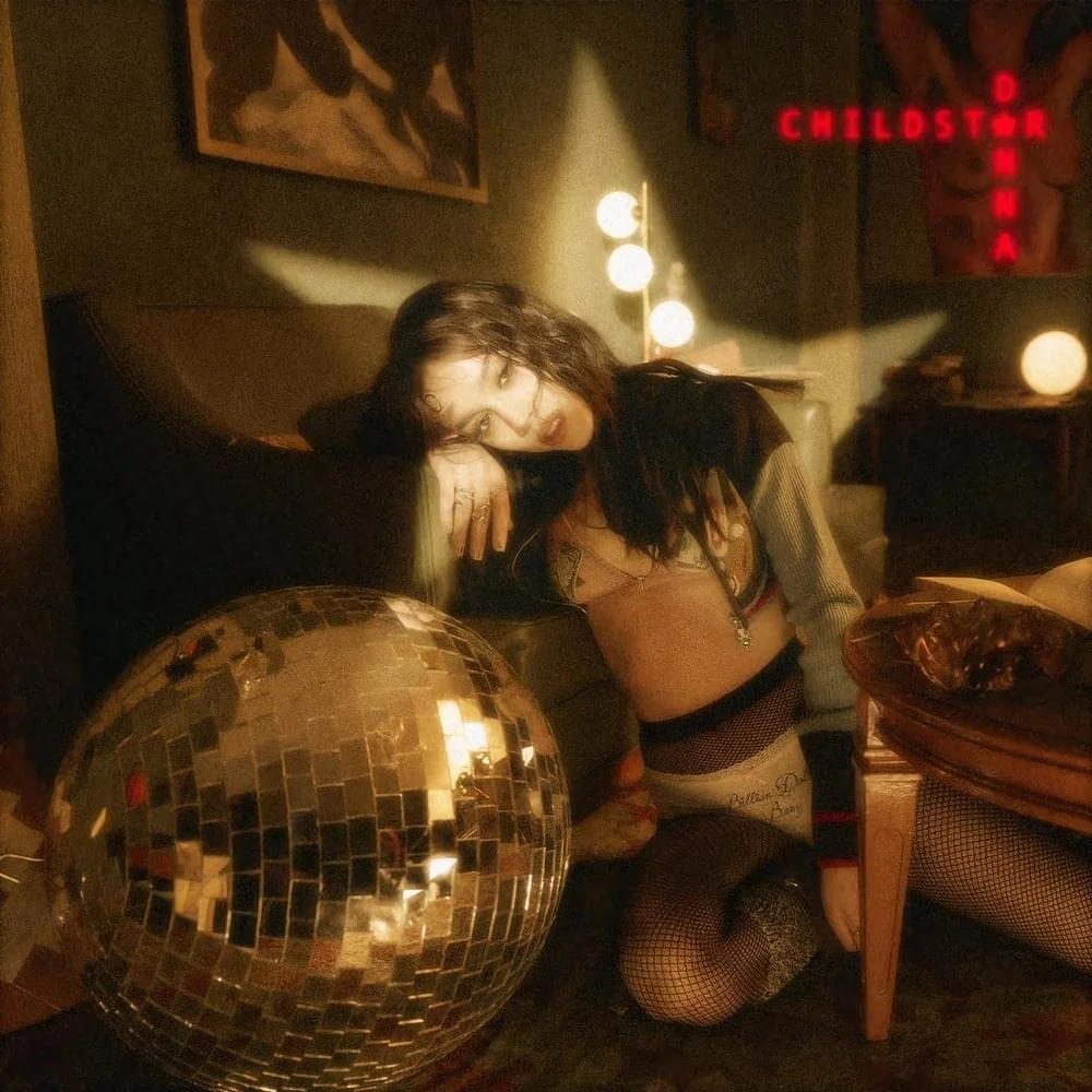 Album cover artwork for Danna's Childstar limited edition orange vinyl photographed by Rafael Arroyo. The composition displays the artist on a dark floor inside a dimly lit vintage interior, resting against a leather couch under localized warm lighti