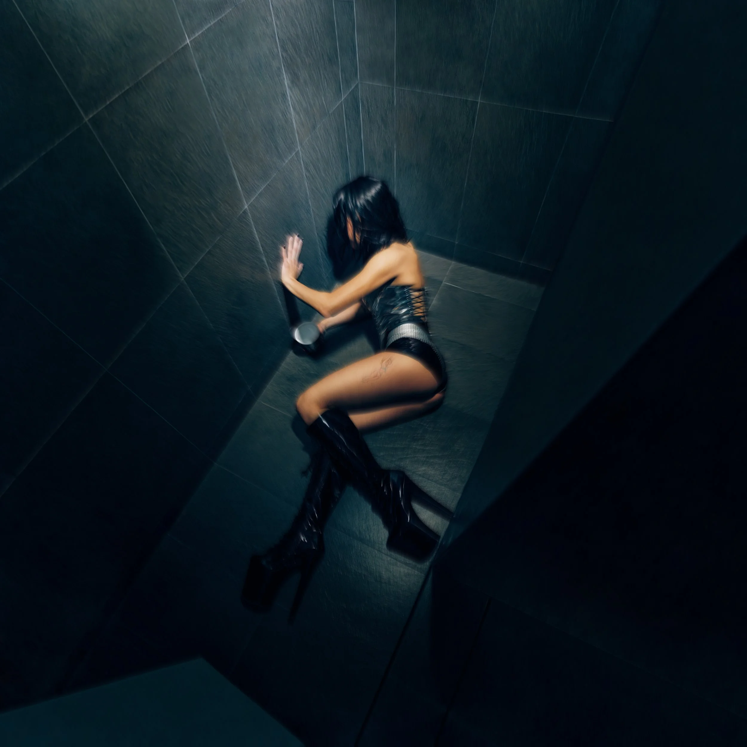 Official single cover for 1trago by Danna, directed and shot by Rafael Arroyo. Top-down view, featuring mexican singer Danna, also known as Danna Paola in a black leather outfit with boots, against a tile floor, leaning into a corner under a bright s