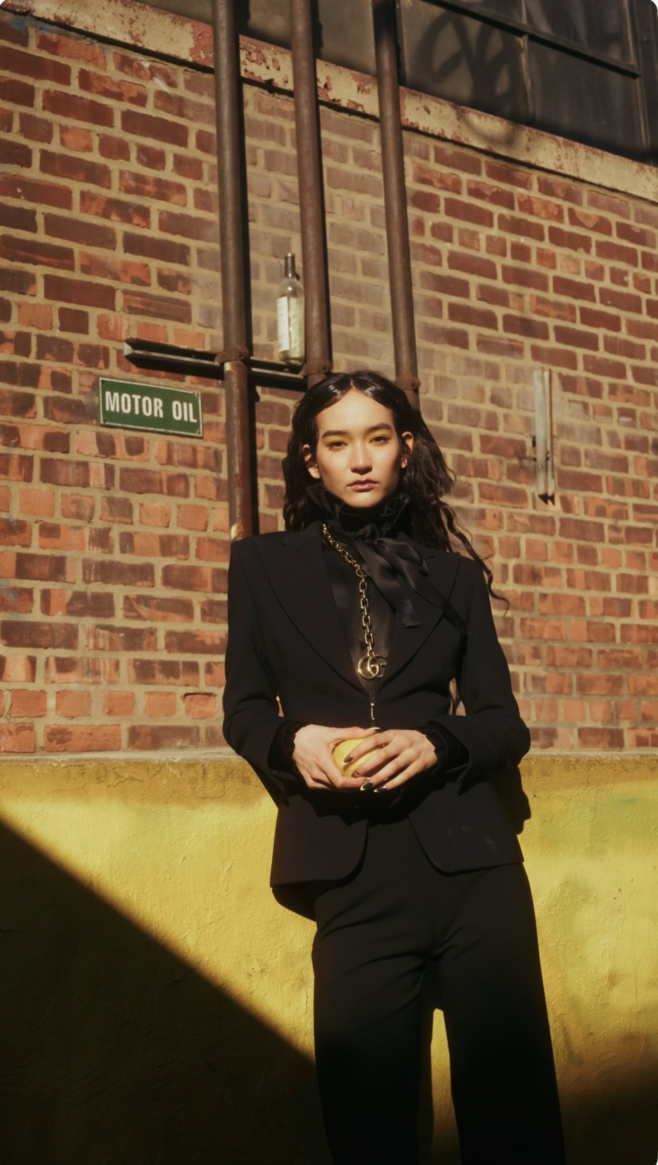 Model in a black Gucci suit stands against a brick wall, in New York, Brooklyn, holding a yellow object with both hands, with a sign that reads 'MOTOR OIL' behind her shot by Rafael Arroyo for the beauty magazine L'Beauté