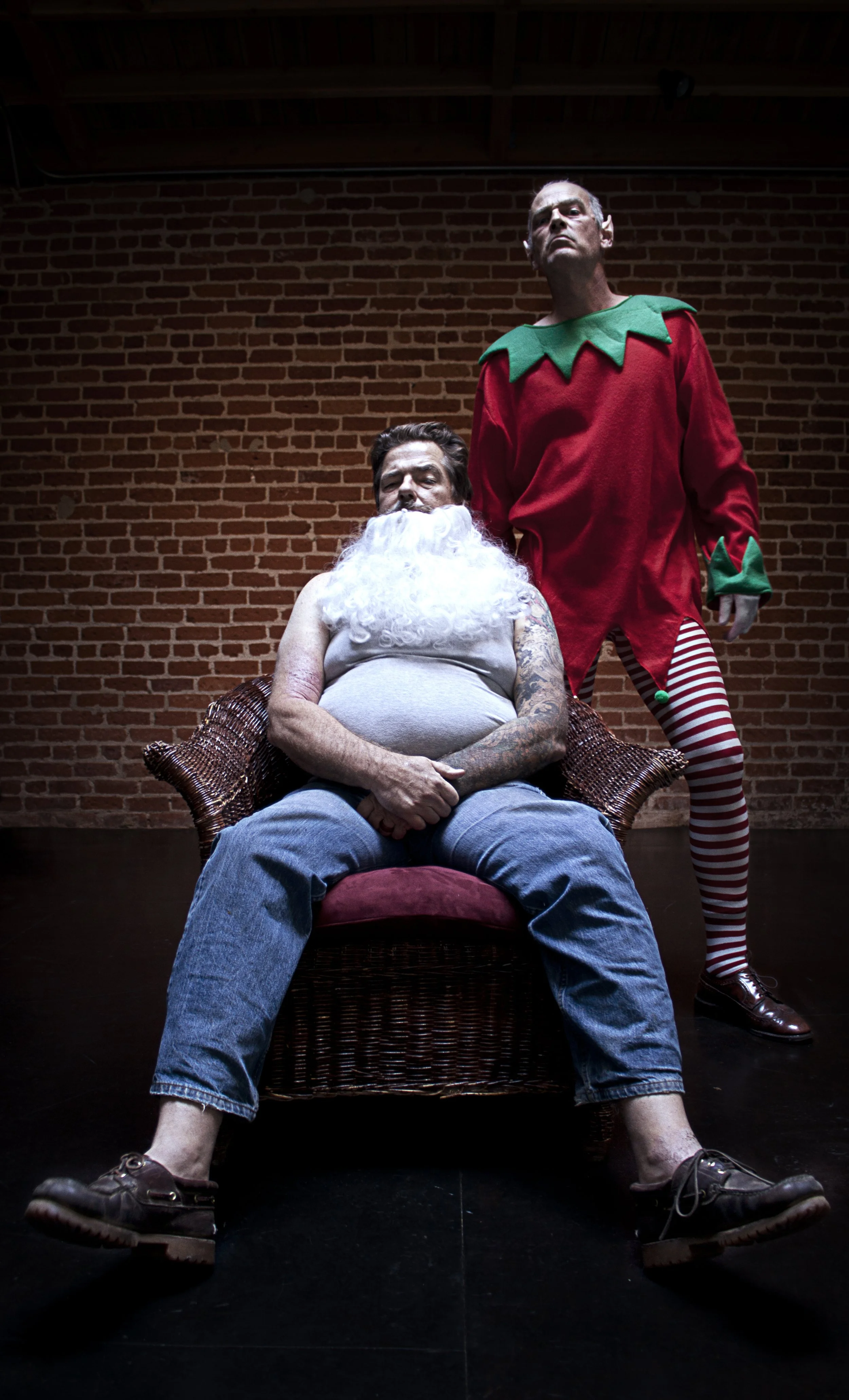 A man sits in a wicker chair with a disheveled beard, wearing a gray tank top and jeans, with tattoos on his arms. Behind him stands a person in an elf costume with a red shirt and striped tights, with a serious expression, against a brick wall backg