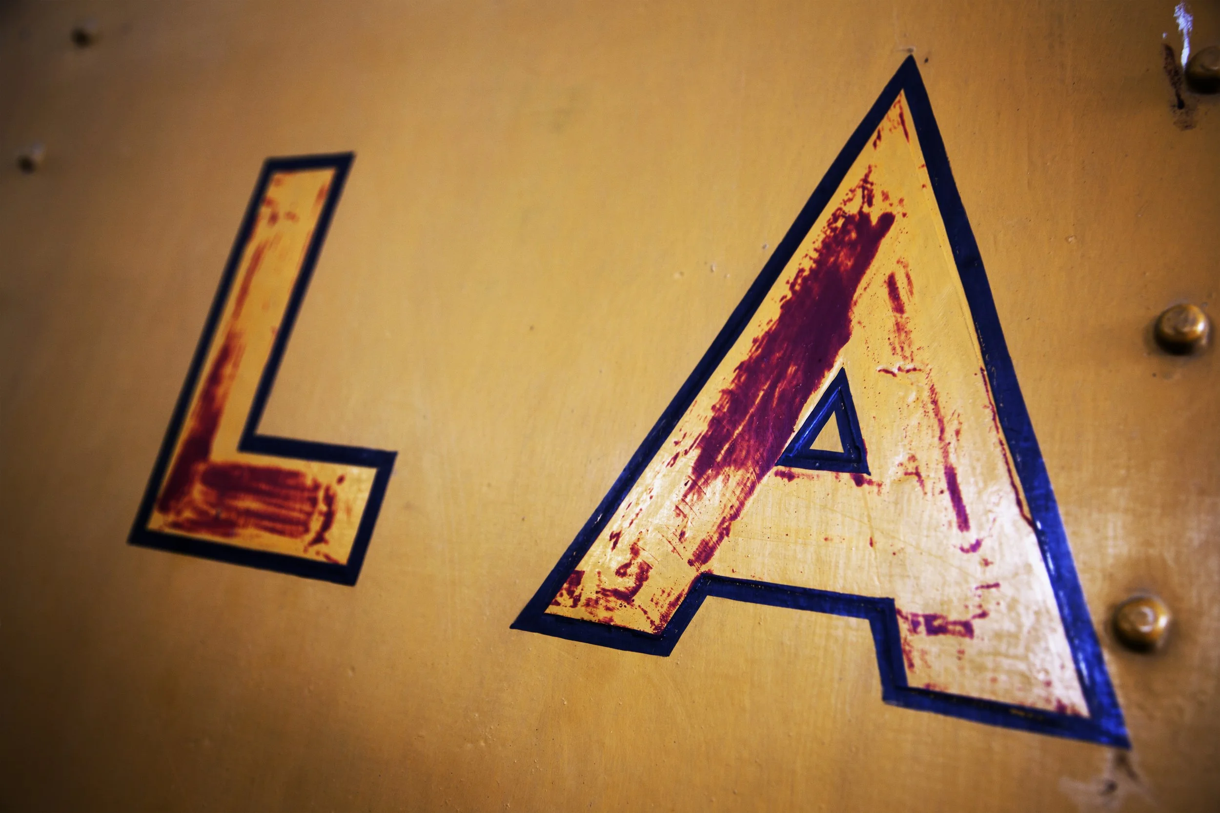Close-up of a yellow sign with a black outline of a large letter 'LA', showing rust and wear, attached to a metal surface with visible screws.