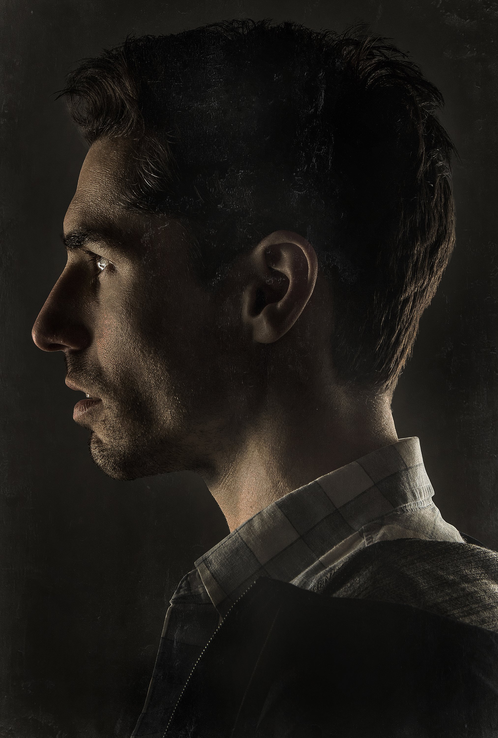 Side profile of a man with dark hair wearing a checkered shirt and a dark jacket, against a black background.
