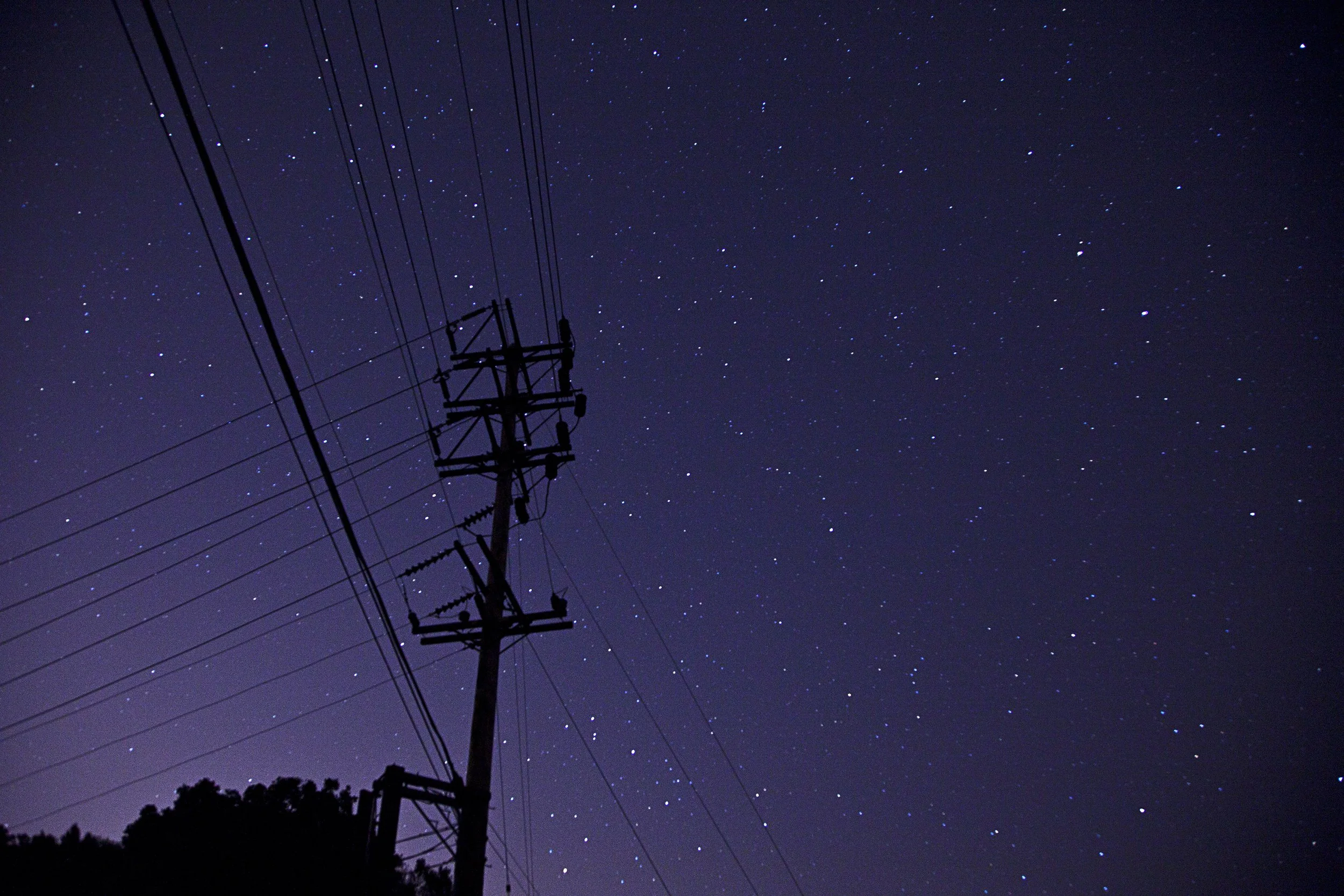 A silhouette of an electricity pole and power lines against a starry night sky.