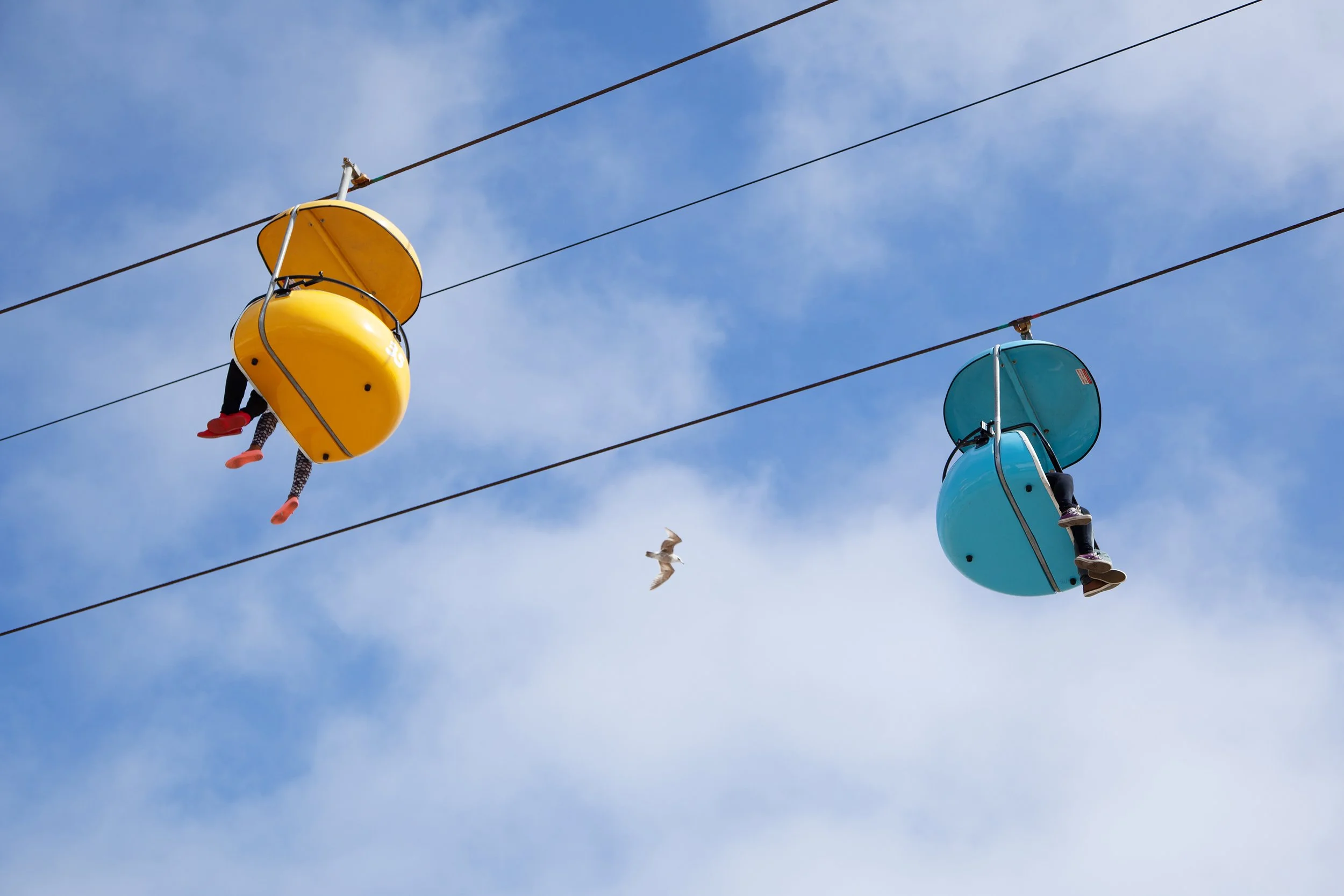 Colorful people riding a zip line high in the sky with a clear blue sky and some clouds in the background.