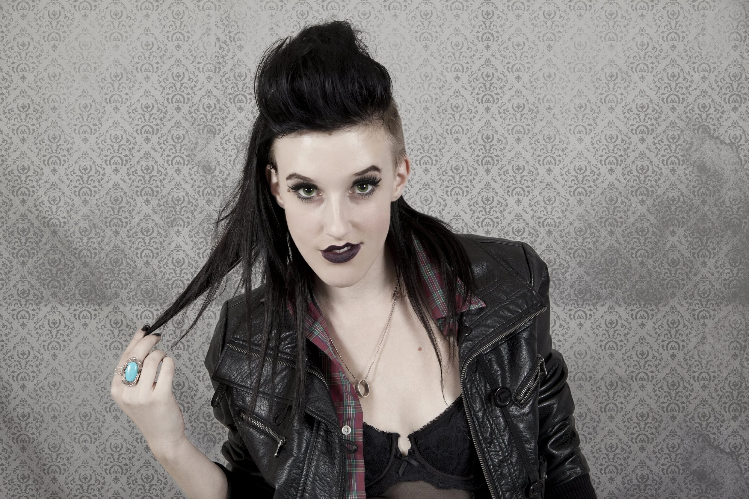 A young woman with dark hair styled in a tall pompadour, green eyes, dark lipstick, wearing a leather jacket and plaid shirt, holding a strand of her hair in front of a gray patterned wall.