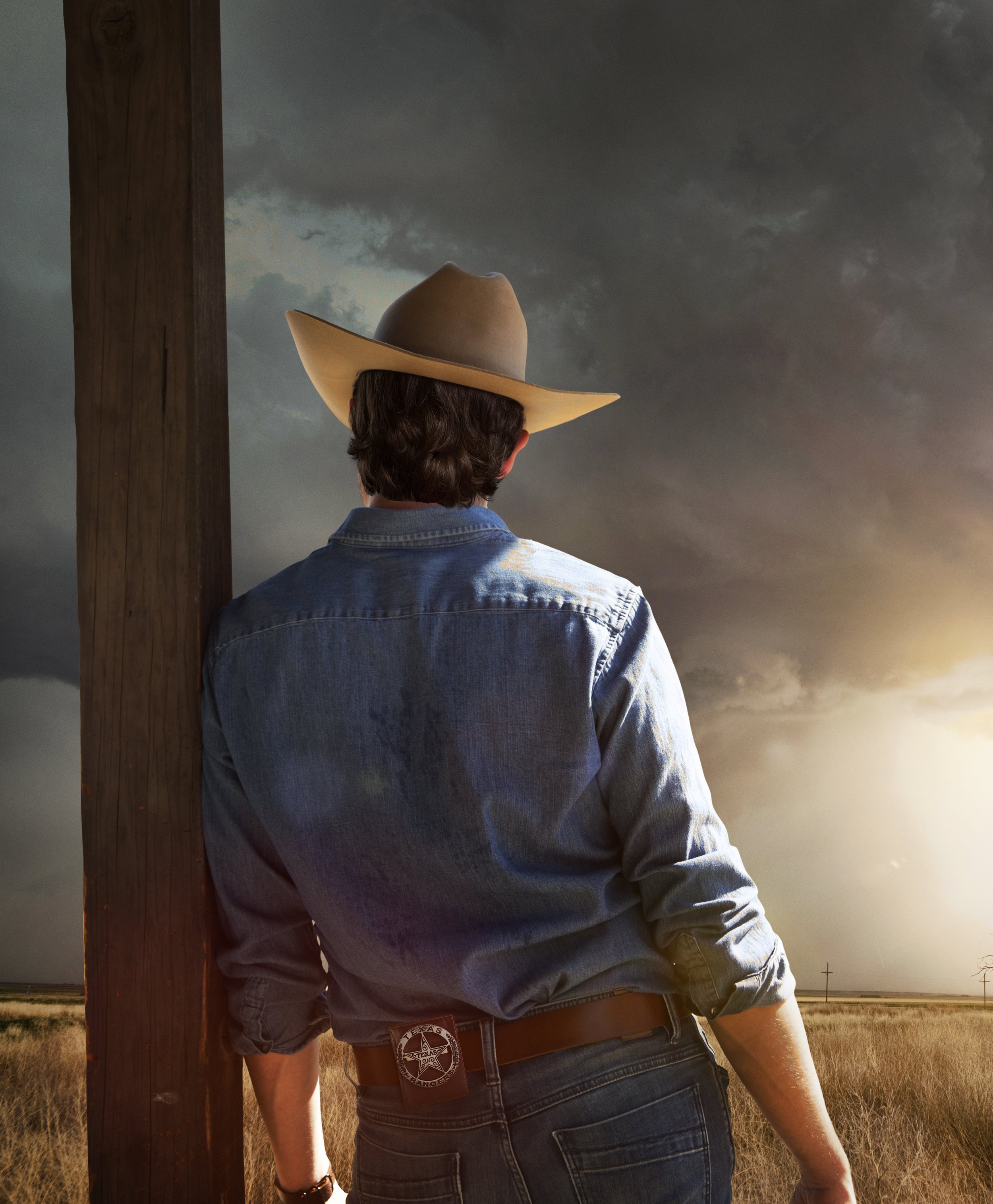 A person wearing a cowboy hat, denim jacket, and dark jeans stands outdoors at sunset, facing away from the camera near a utility pole and an open field with a cloudy sky.