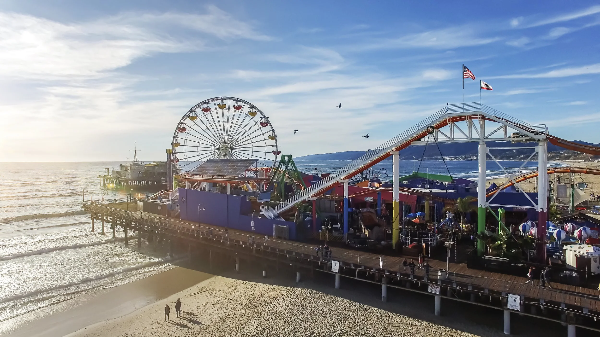 A beachside amusement park with a Ferris wheel and roller coaster, located on a pier extending into the ocean, with a sailboat in the water and a few people walking on the beach.