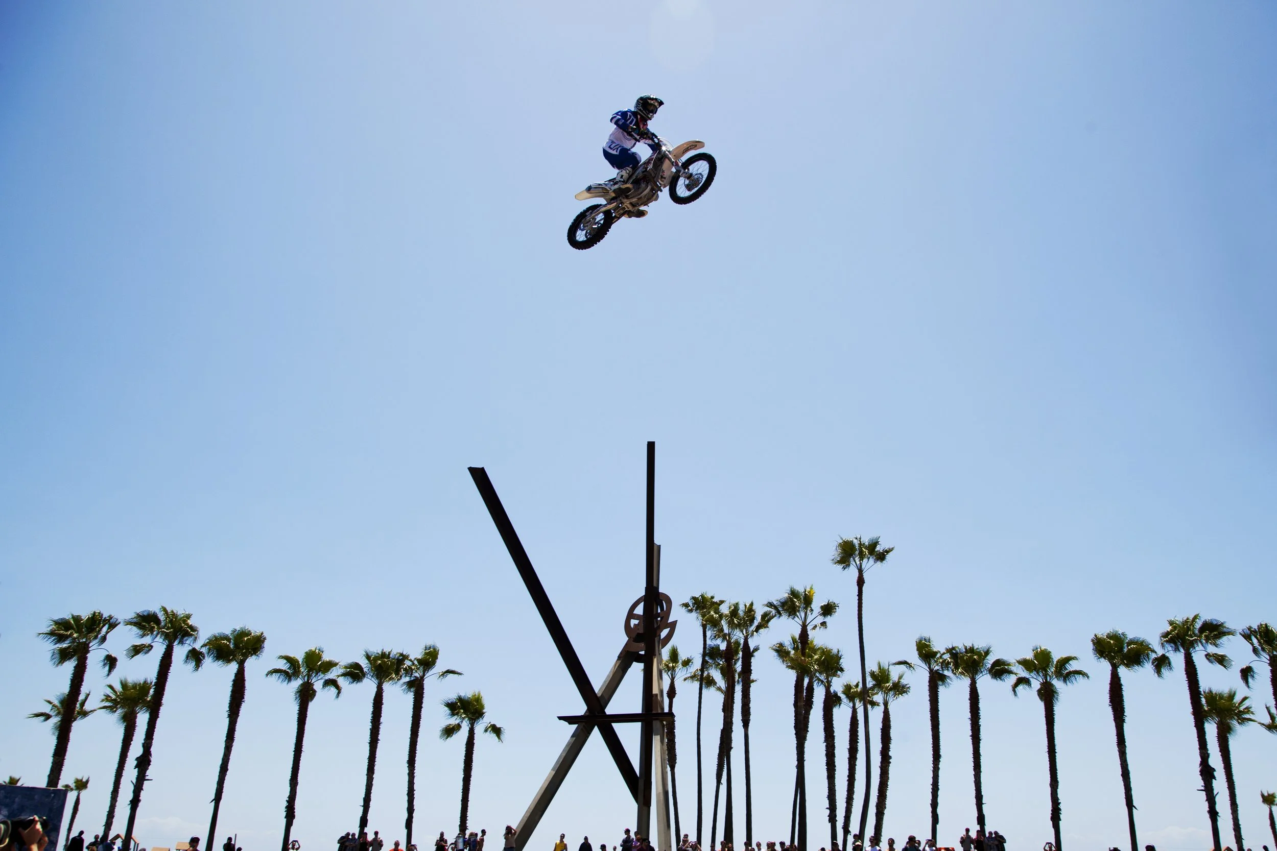 A person riding a motorcycle through the air during a stunt at an outdoor event, with a row of palm trees and a crowd of spectators visible at the bottom, all set against a clear blue sky.