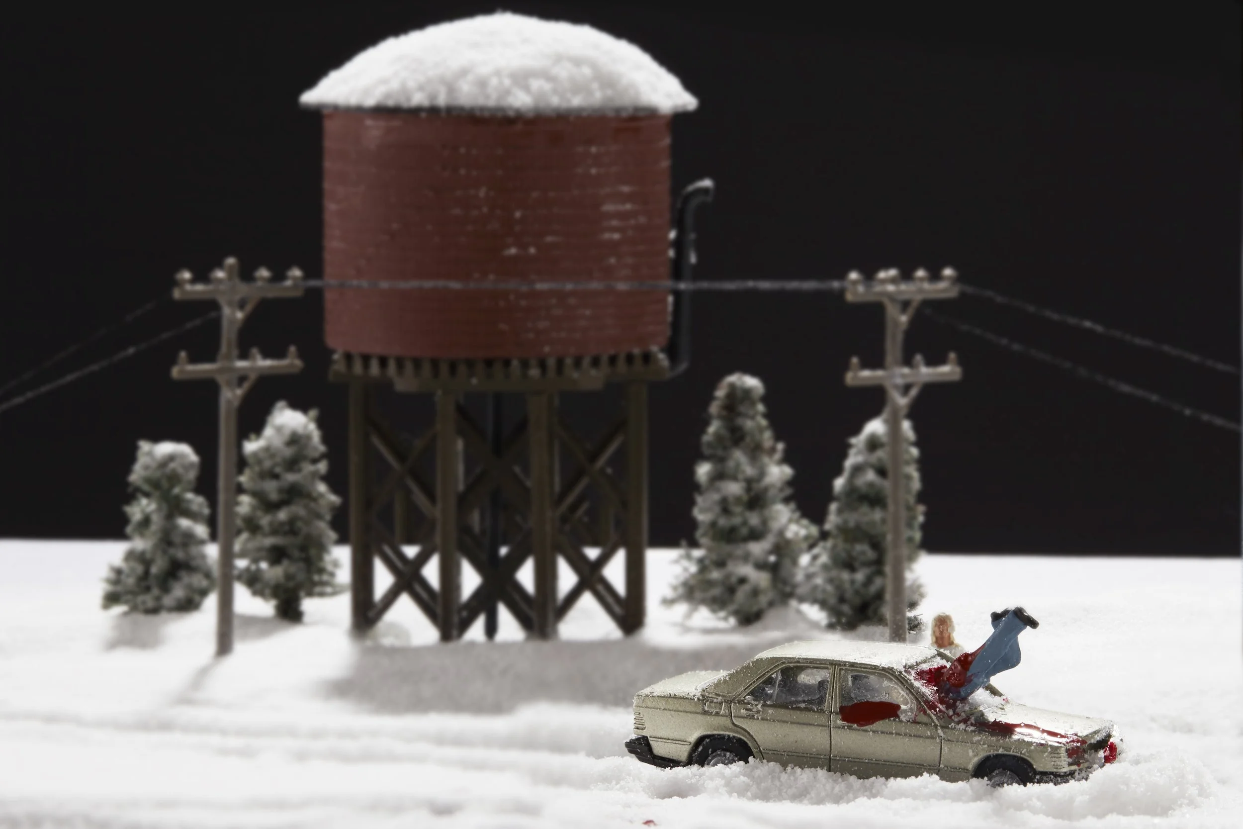 Miniature winter scene featuring a small red water tower with snow on top, surrounded by snow-covered trees, electrical poles, and wires, with a toy car partially covered in snow and a small toy figure lying face down in the snow.