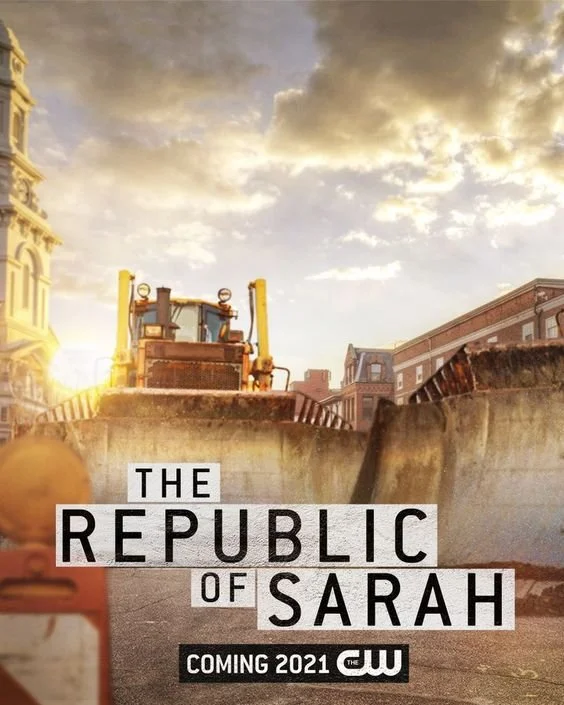 A bulldozer parked on a city street during sunset, with buildings on both sides and a cloudy sky.