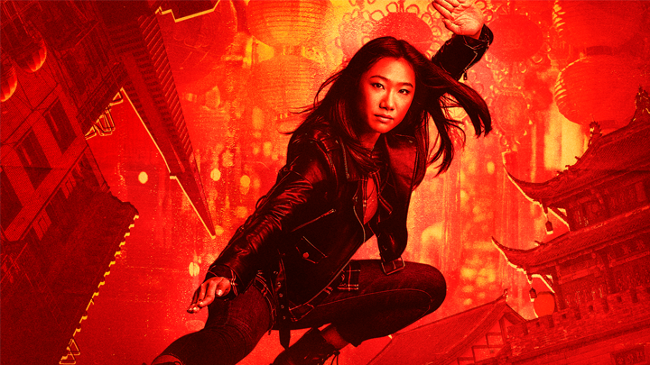 A young woman in a black leather jacket and black jeans crouches in an urban setting with traditional Asian architecture. The background is vibrant red with lanterns and festive decorations.
