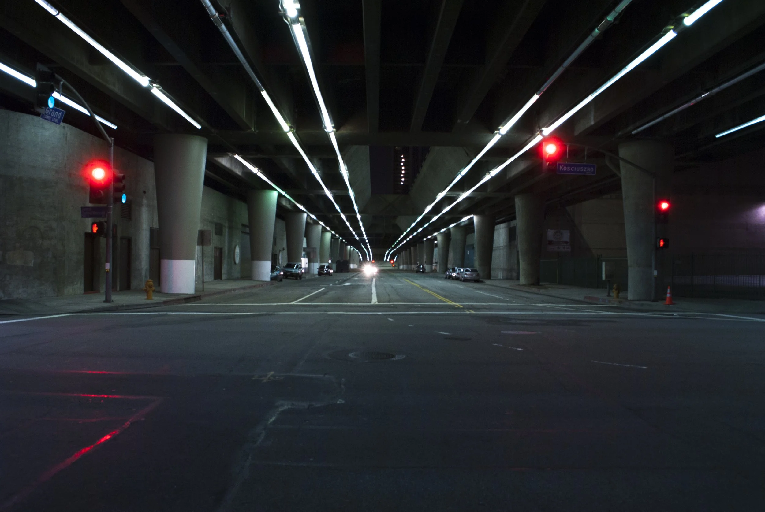 Night view of an urban underpass with lit overhead pipes, traffic lights, and cars parked along the sides.