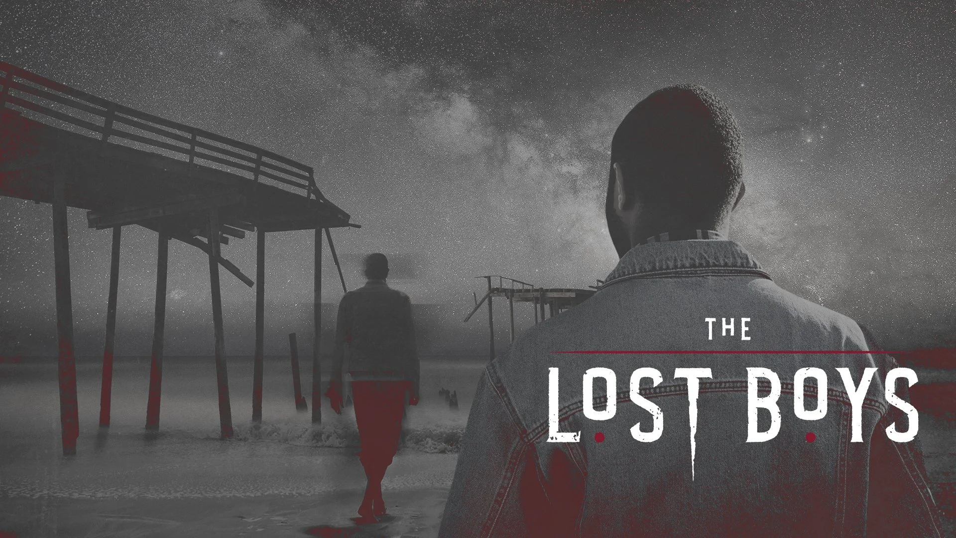 Black and white photo of three young men standing on a beach at night, looking up at a starry sky with the Milky Way visible. The man in the foreground has short hair and is wearing a denim jacket, with the title 'The Lost Boys' partially visible in 