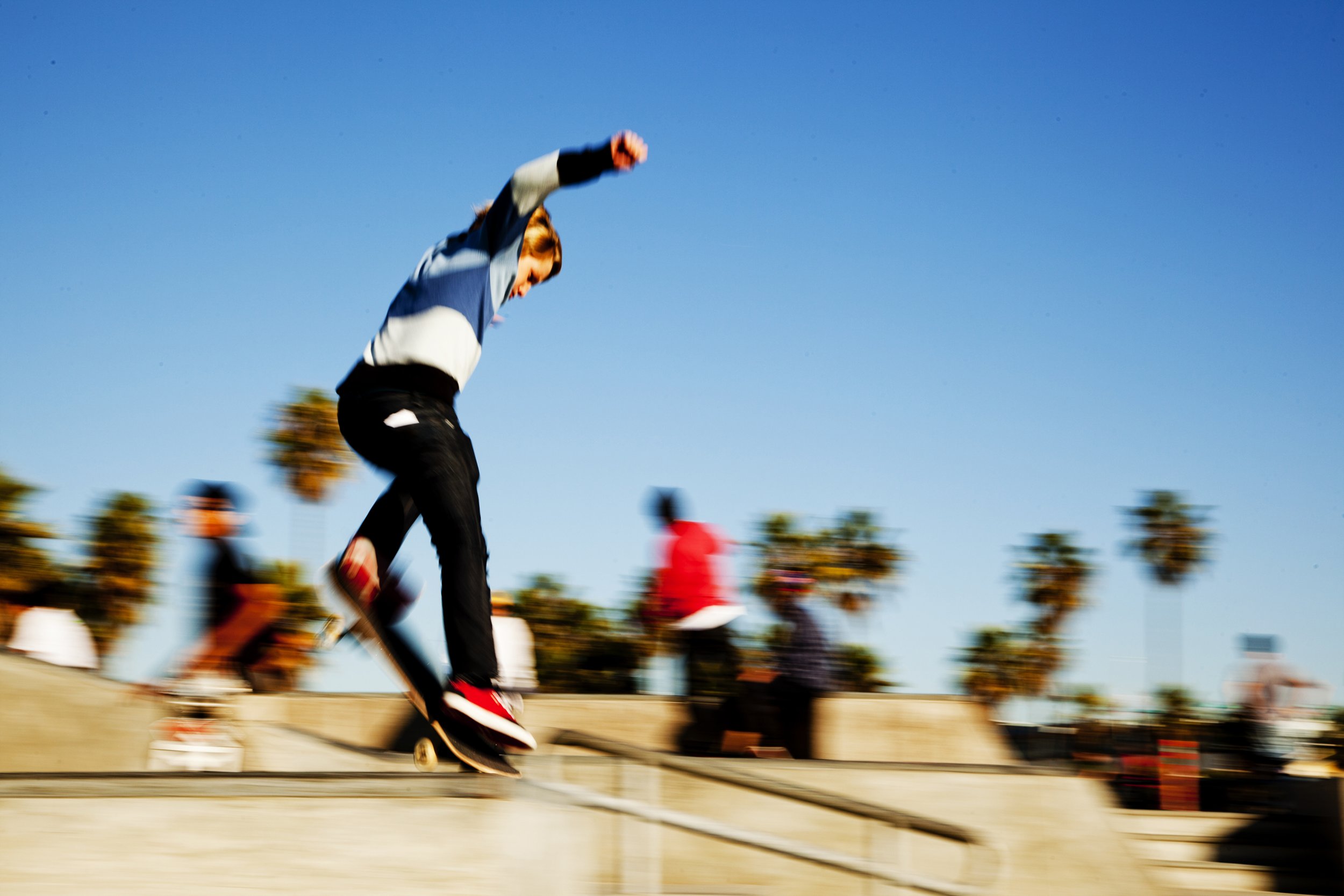 A person riding a skateboard at a skate park with blurred onlookers and palm trees in the background under a clear blue sky.