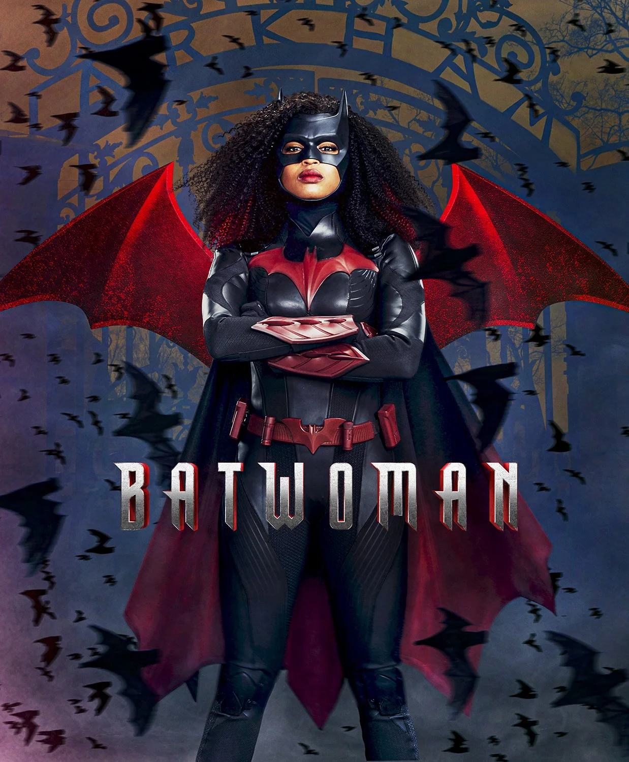 A person dressed as Batwoman in a superhero costume with red and black armor, red cape, and bat mask, standing with arms crossed in front of a spooky background with bats and a wrought iron gate.