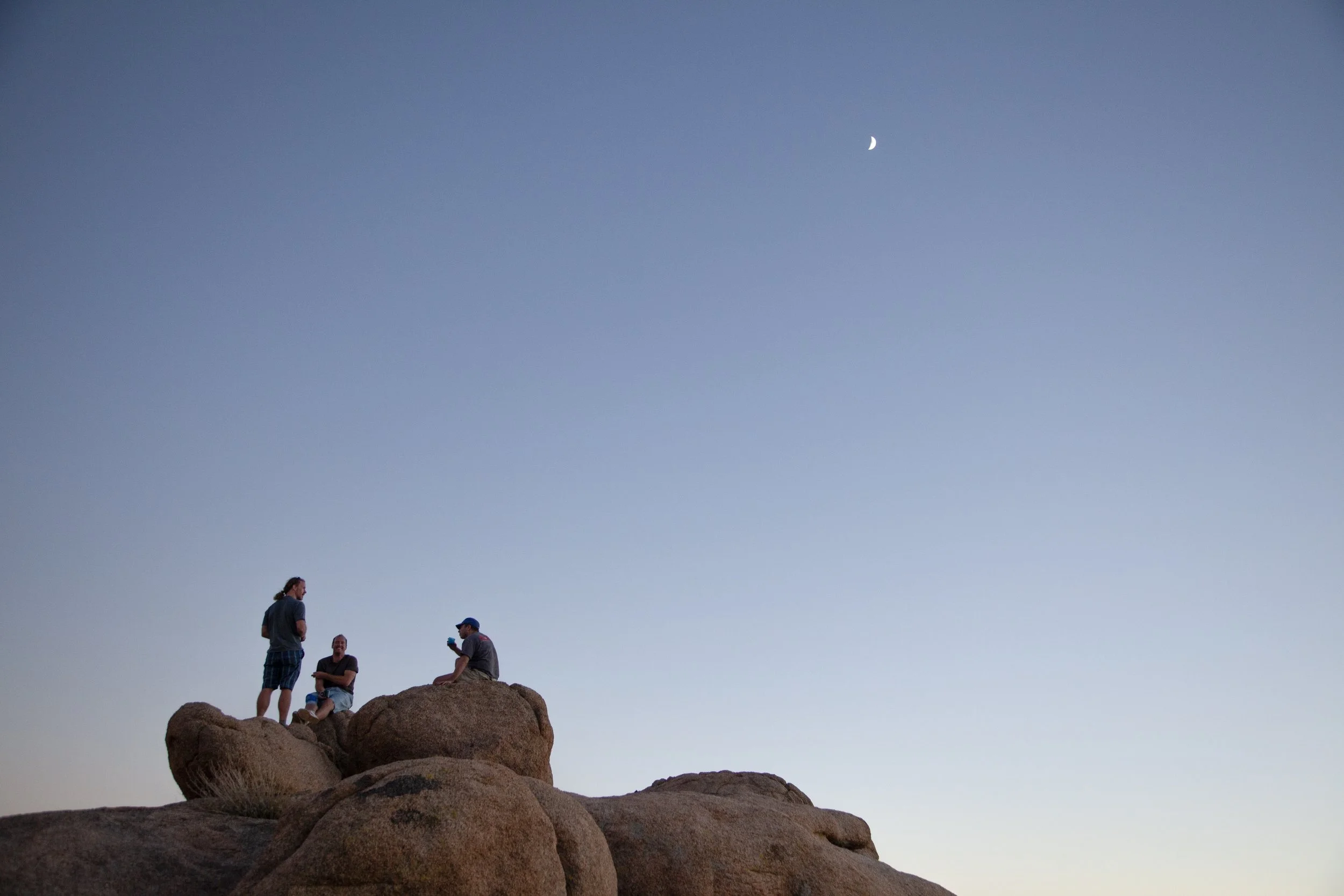 Four people standing and sitting on large rocks outdoors at dusk with a clear sky and a visible crescent moon.