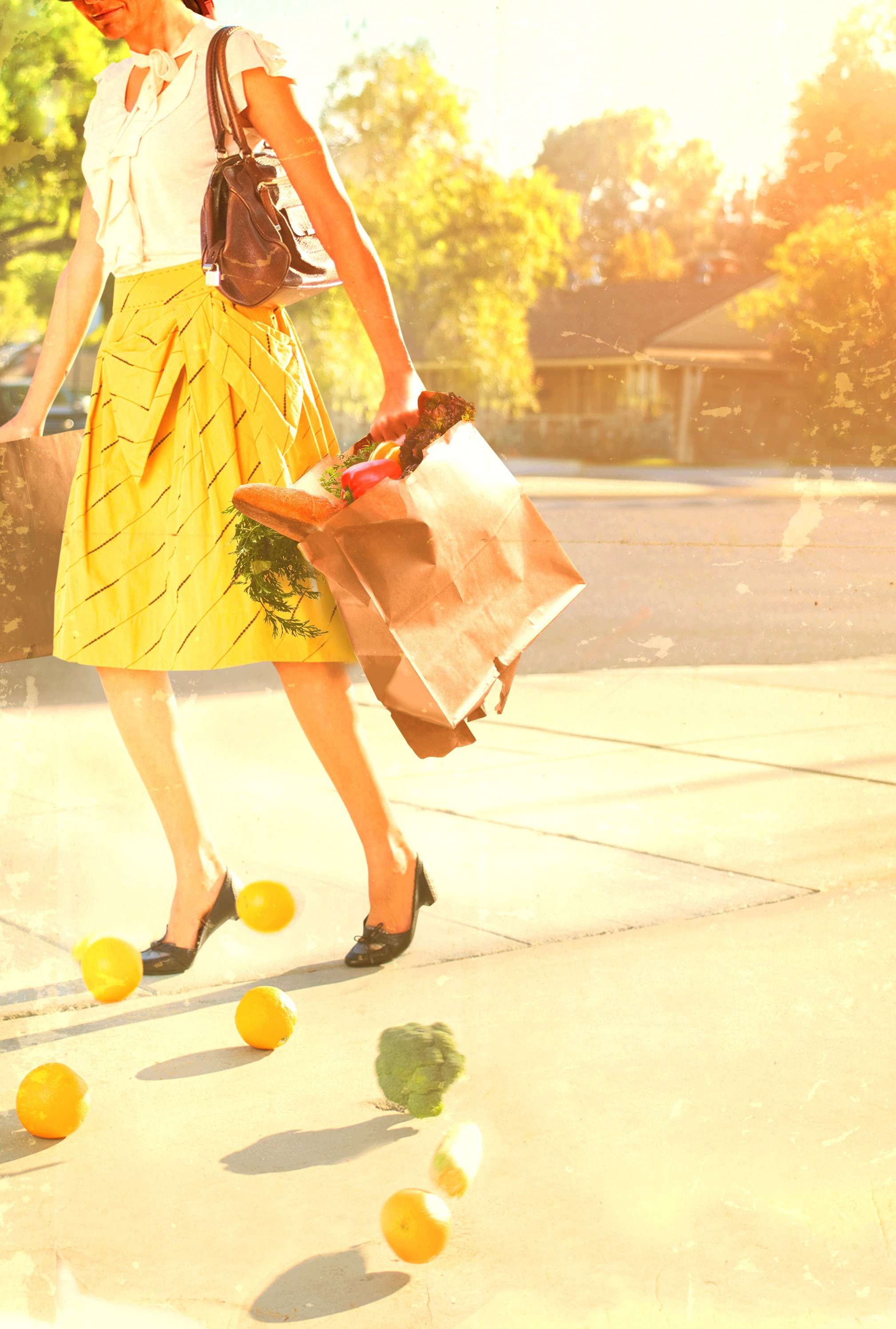 A woman in a yellow skirt and black shoes is walking outdoors on a sunny day, carrying grocery bags with vegetables, with oranges and broccoli on the ground.