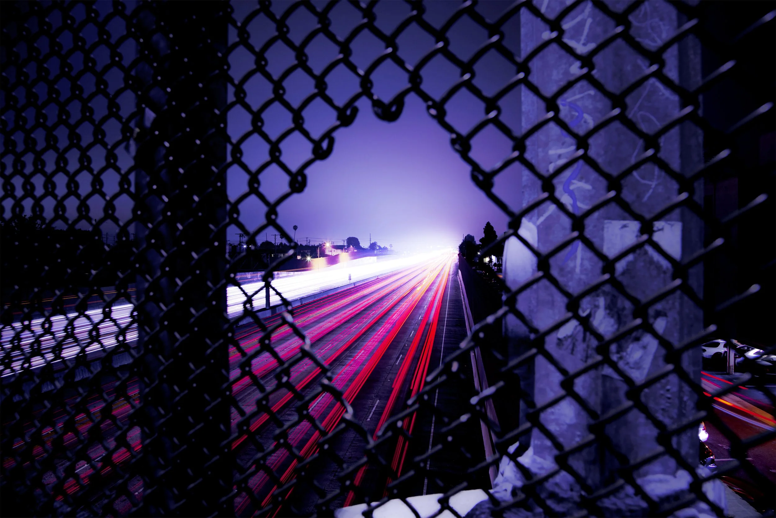 Nighttime city scene with light trails on a highway seen through a chain-link fence, illuminated by purple and blue lights.