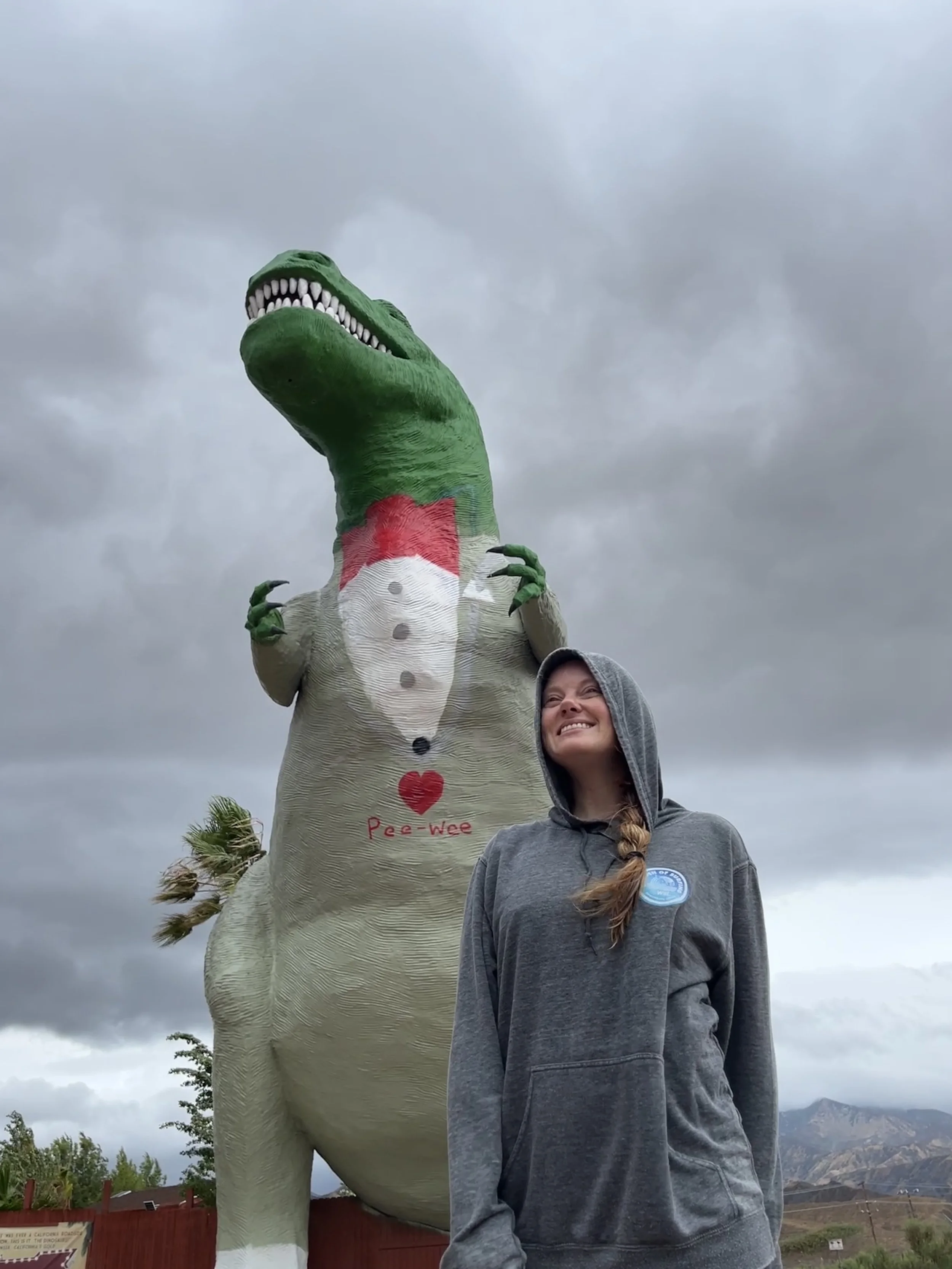 A woman with long hair in a braid, wearing a gray hoodie, smiling and standing next to a large dinosaur statue with a green head and white body, under a cloudy sky.