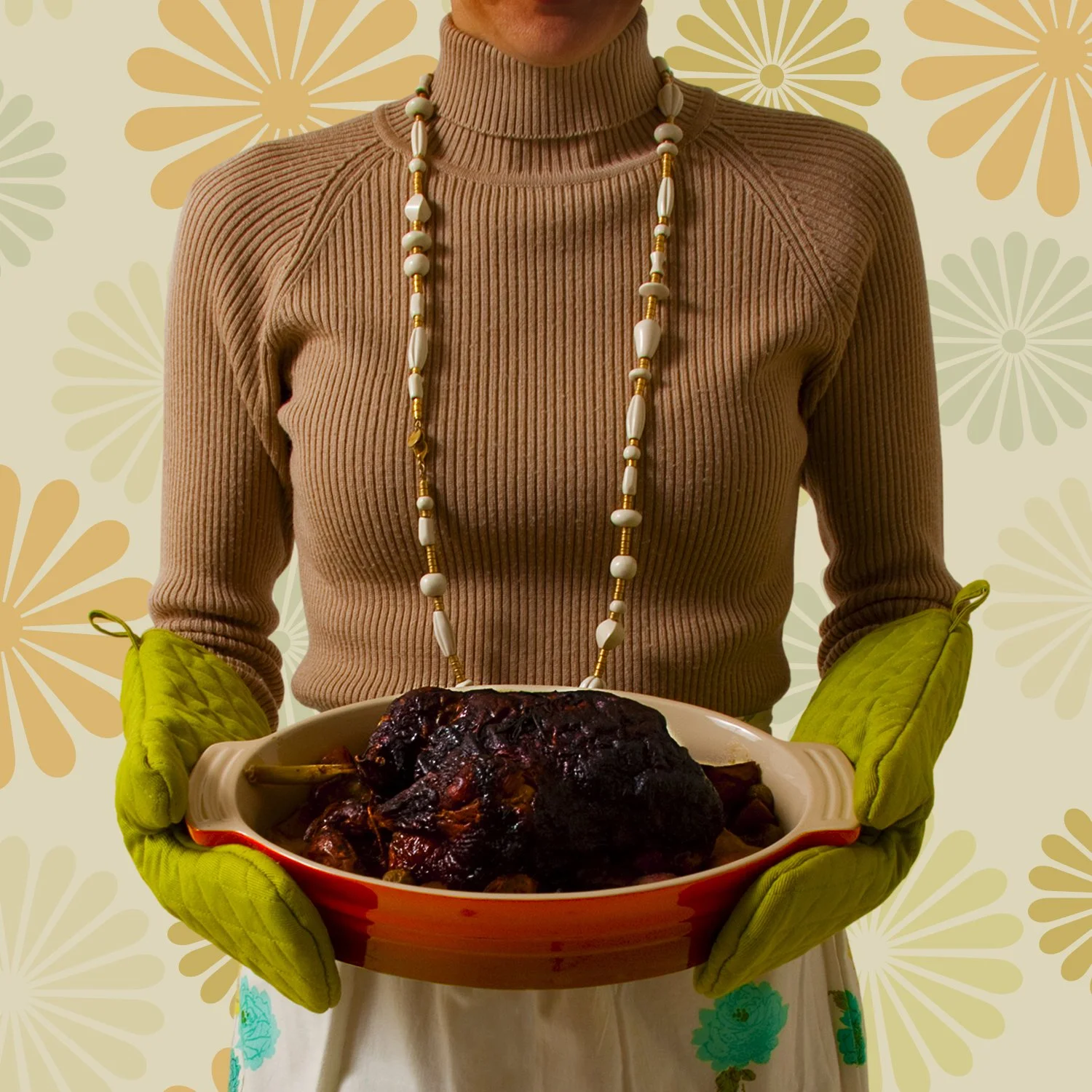 Person wearing a beige turtleneck sweater and long beaded necklace holding a dish with a large, cooked, dark-colored meat, wearing green oven mitts, in front of a floral patterned background.